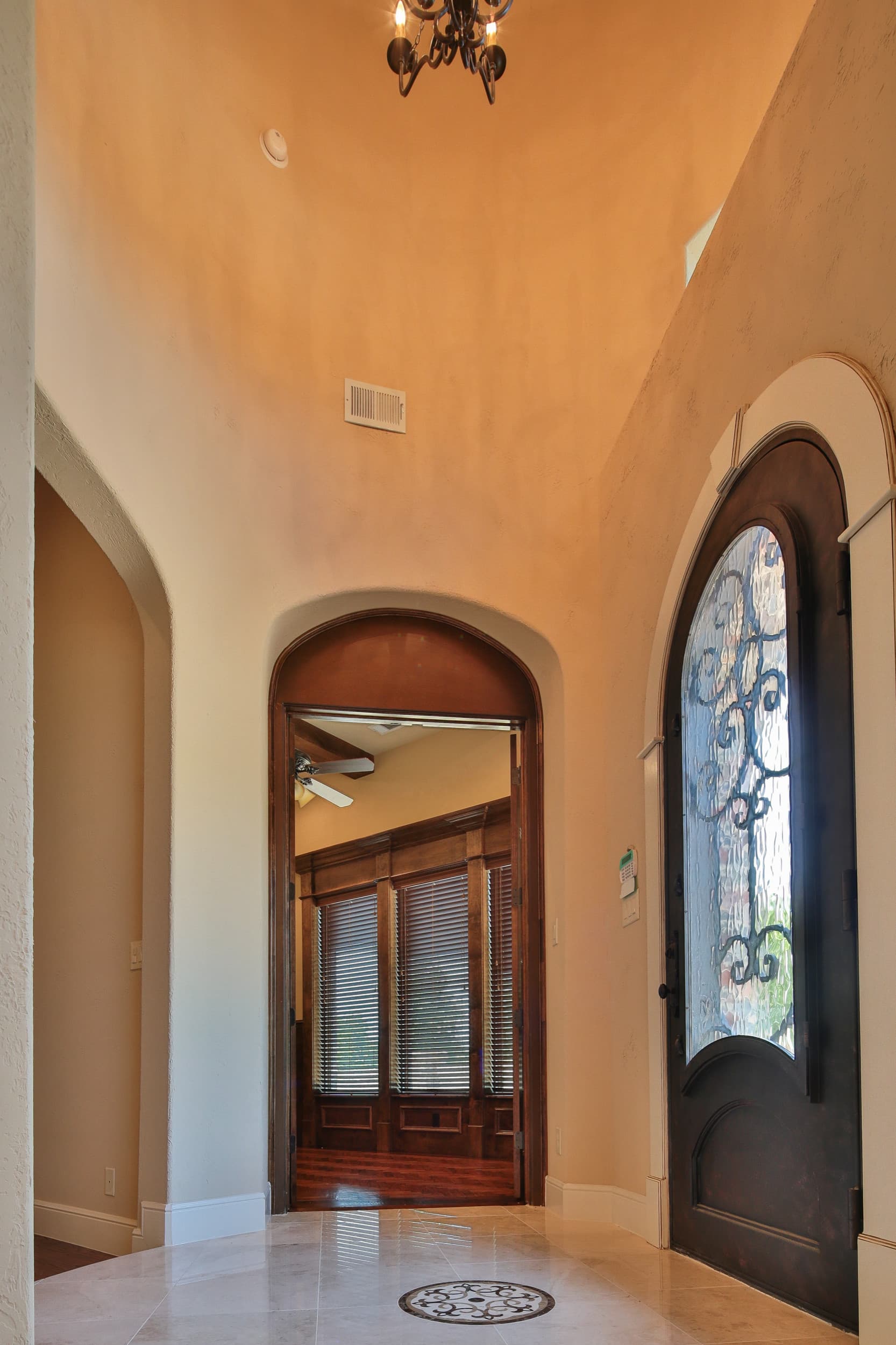 Curved foyer with iron chandelier, stone tile floor with inlaid medallion, arched iron-and-glass front door, and arched opening to a wood-paneled study