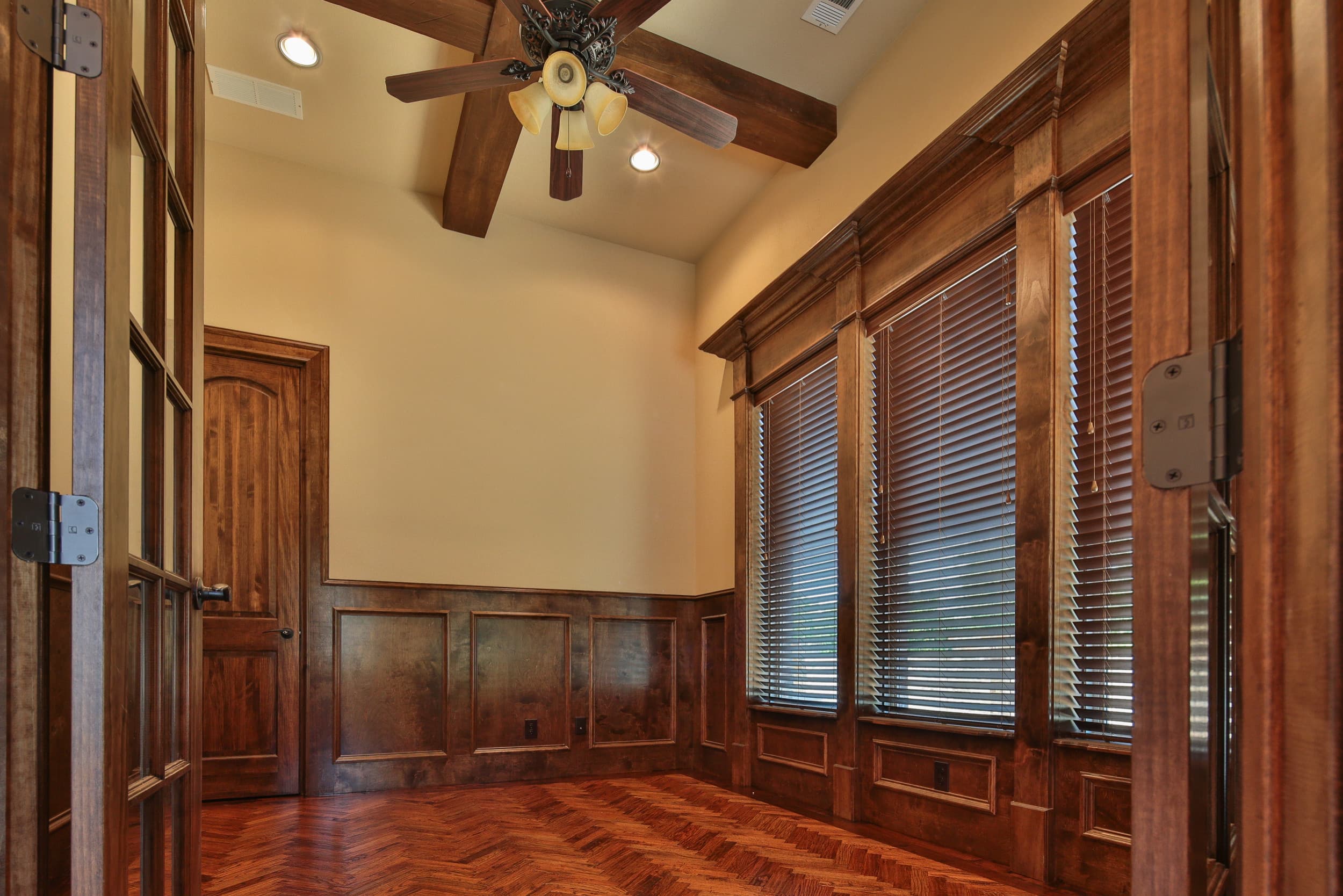 Study framed by open French doors, with stained wood wainscoting, herringbone hardwood floor, wood ceiling beams, ceiling fan, and tall windows