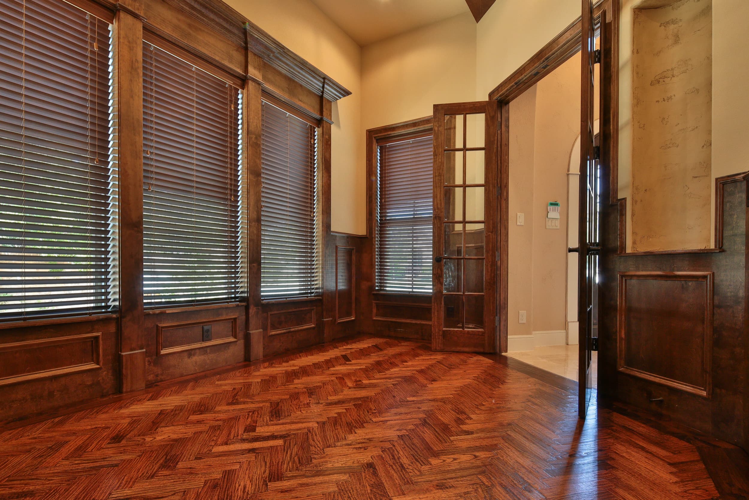 Study with herringbone hardwood floor, stained wood wainscot paneling, tall windows with wood blinds, and open glass-paned French doors to the foyer