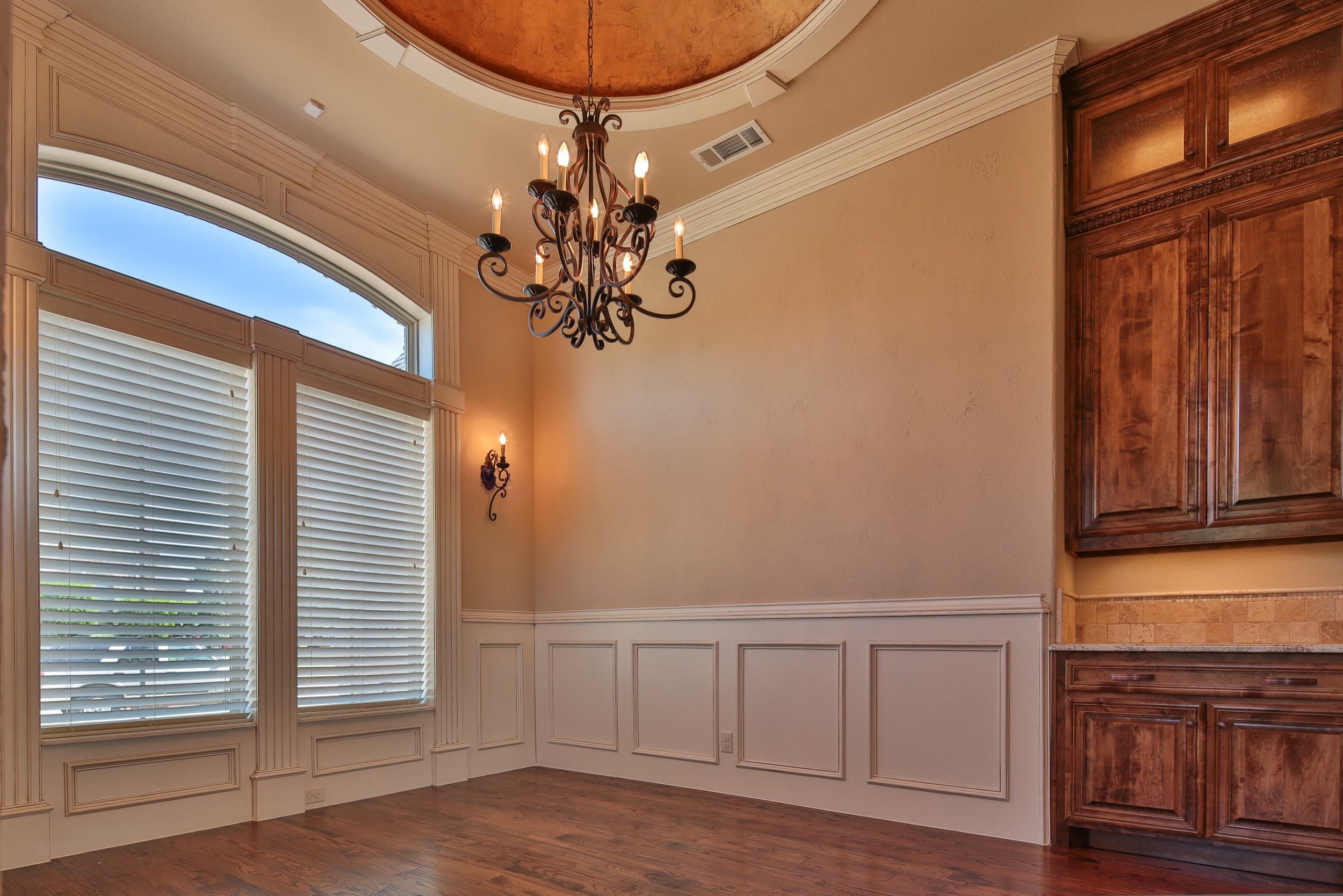 Dining room with tall windows under an arched transom, wrought iron chandelier, white wainscoting, hardwood floor, and stained wood built-in buffet