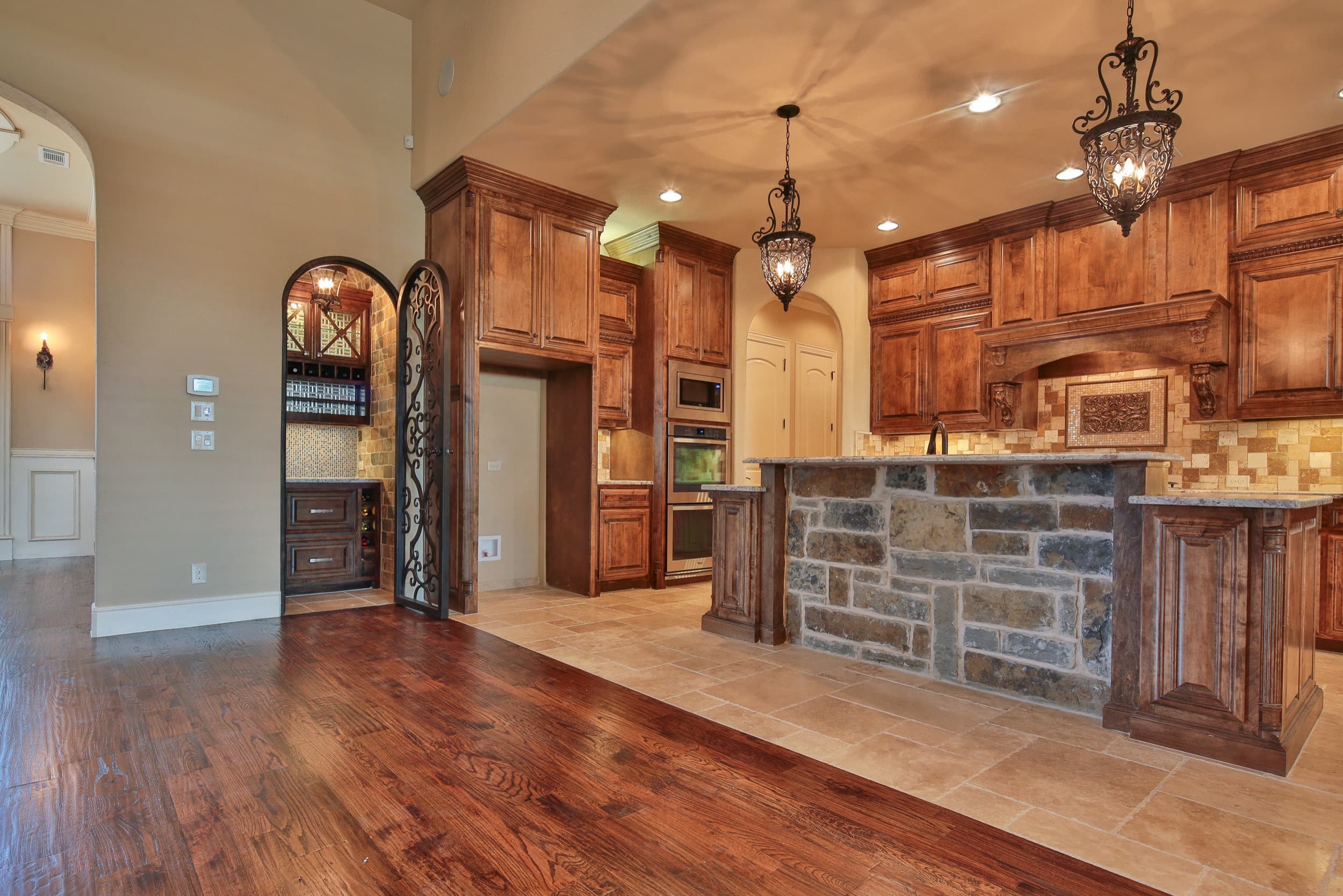 Kitchen with stained wood cabinetry, natural stone island base, granite counters, iron lantern pendants, travertine backsplash and floor, and stainless wall ovens