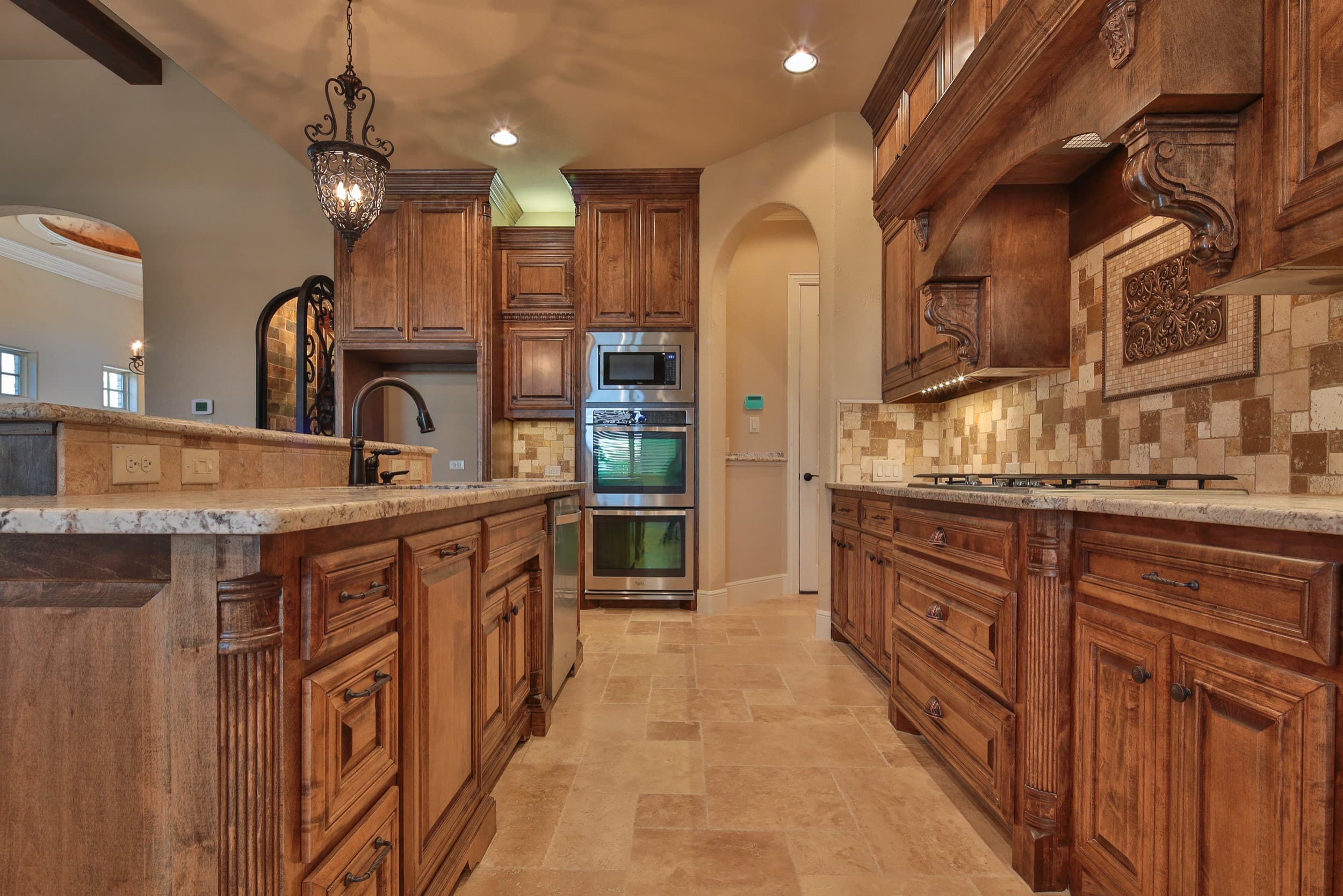 Kitchen aisle between a granite-topped island and gas cooktop wall, with stained wood cabinetry, carved corbels, travertine backsplash, and stainless wall ovens