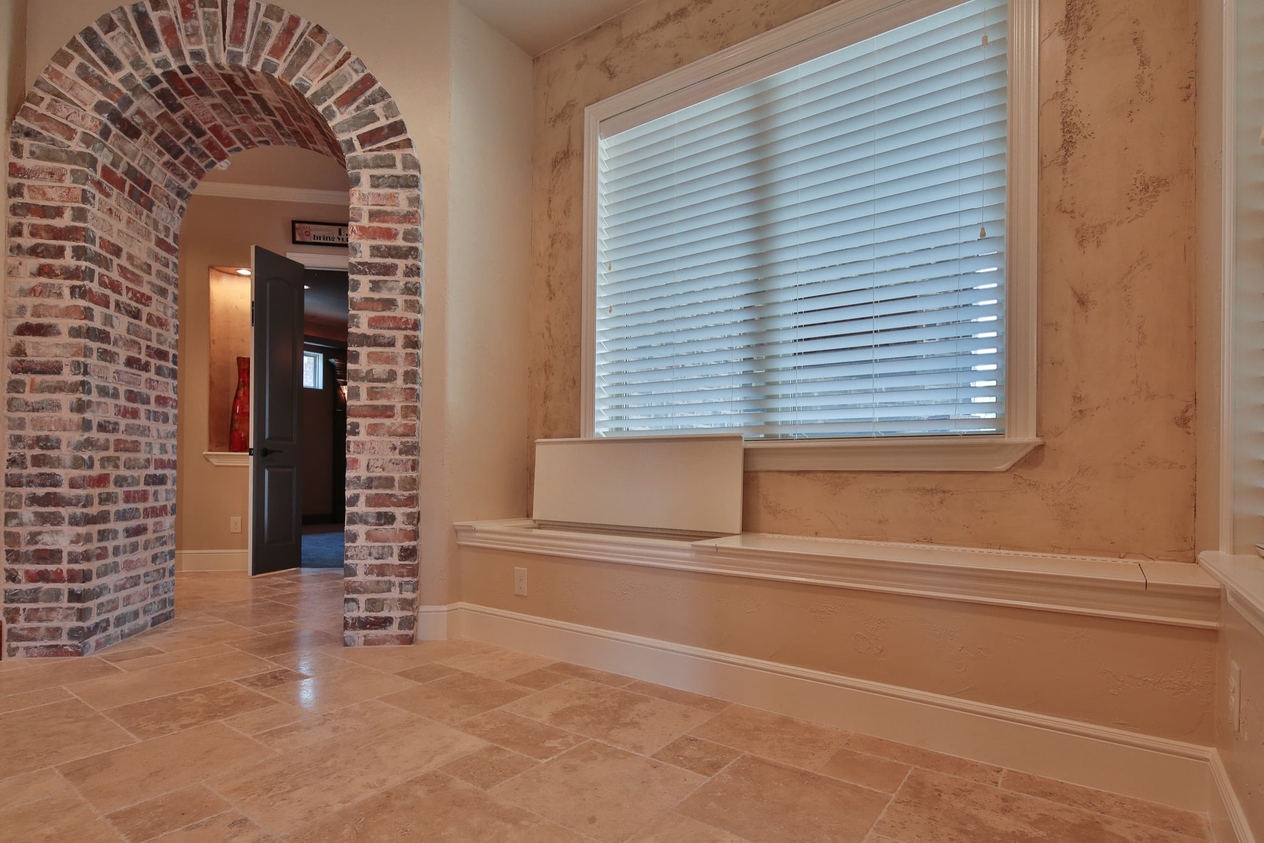 Built-in window seat below a window with white blinds, travertine floor, textured plaster walls, and a brick archway leading to a hall