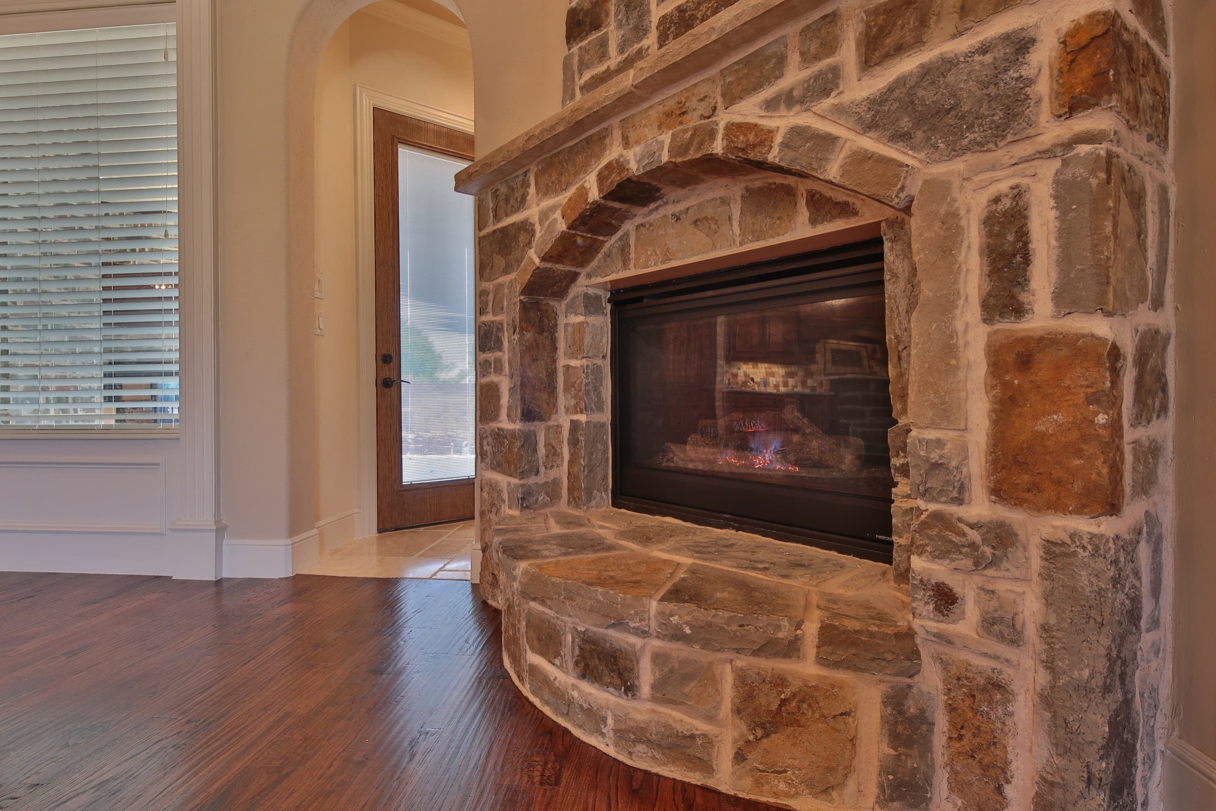 Lit natural stone gas fireplace with arched opening, stone mantel ledge, and curved raised hearth, set on hand-scraped hardwood flooring