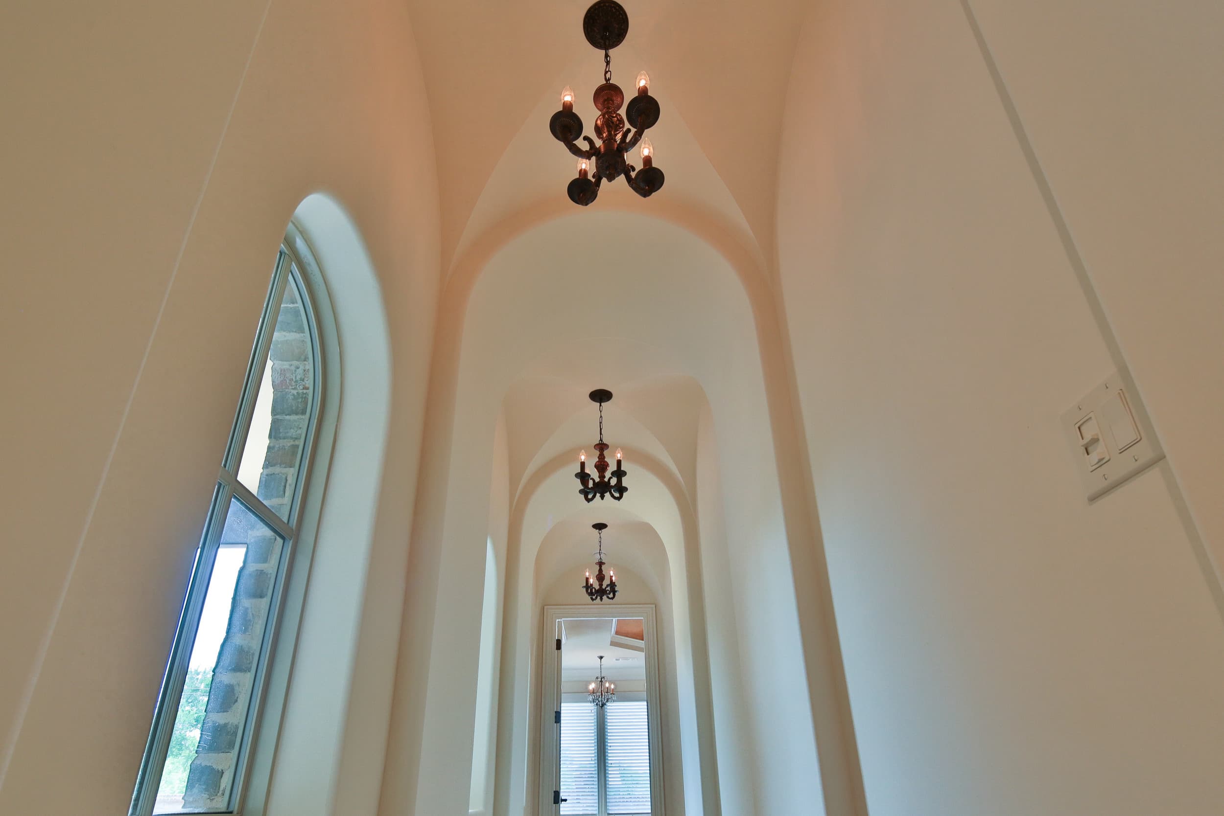 Low-angle view of a groin-vaulted hallway with successive arches, three small bronze candle chandeliers, and a tall arched window