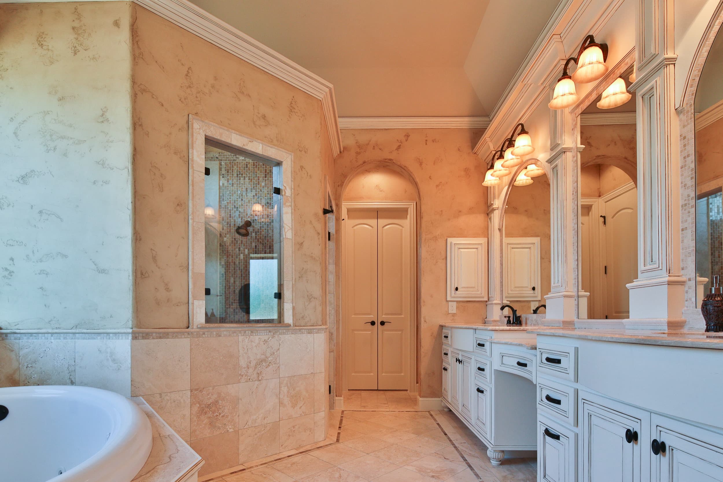 Primary bath with white glazed dual vanities, arched mirrors, bronze sconces, jetted tub, mosaic-tiled walk-in shower, and travertine floor with inlaid border
