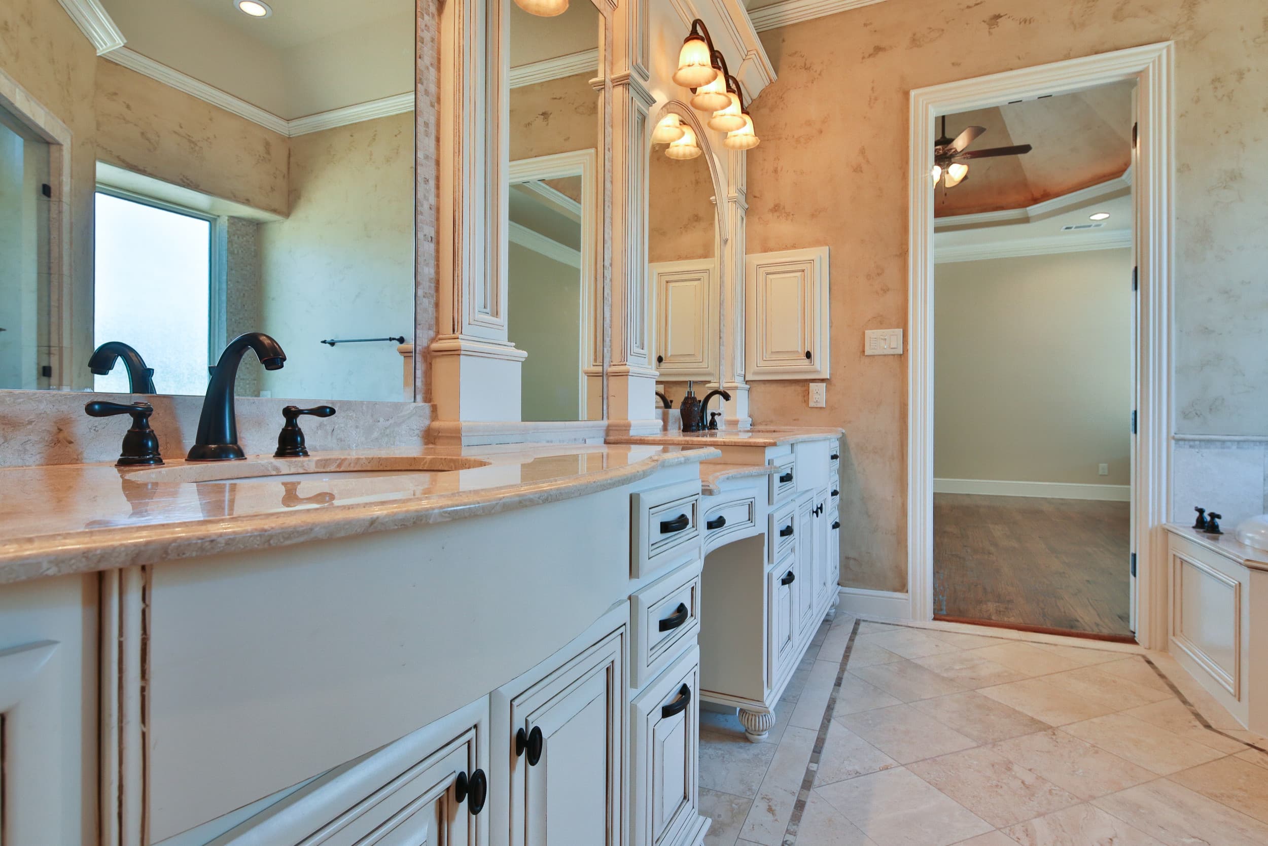 Primary bath with curved white glazed vanity, cream stone counters, oil-rubbed bronze faucets, travertine floor, and an open doorway to the bedroom