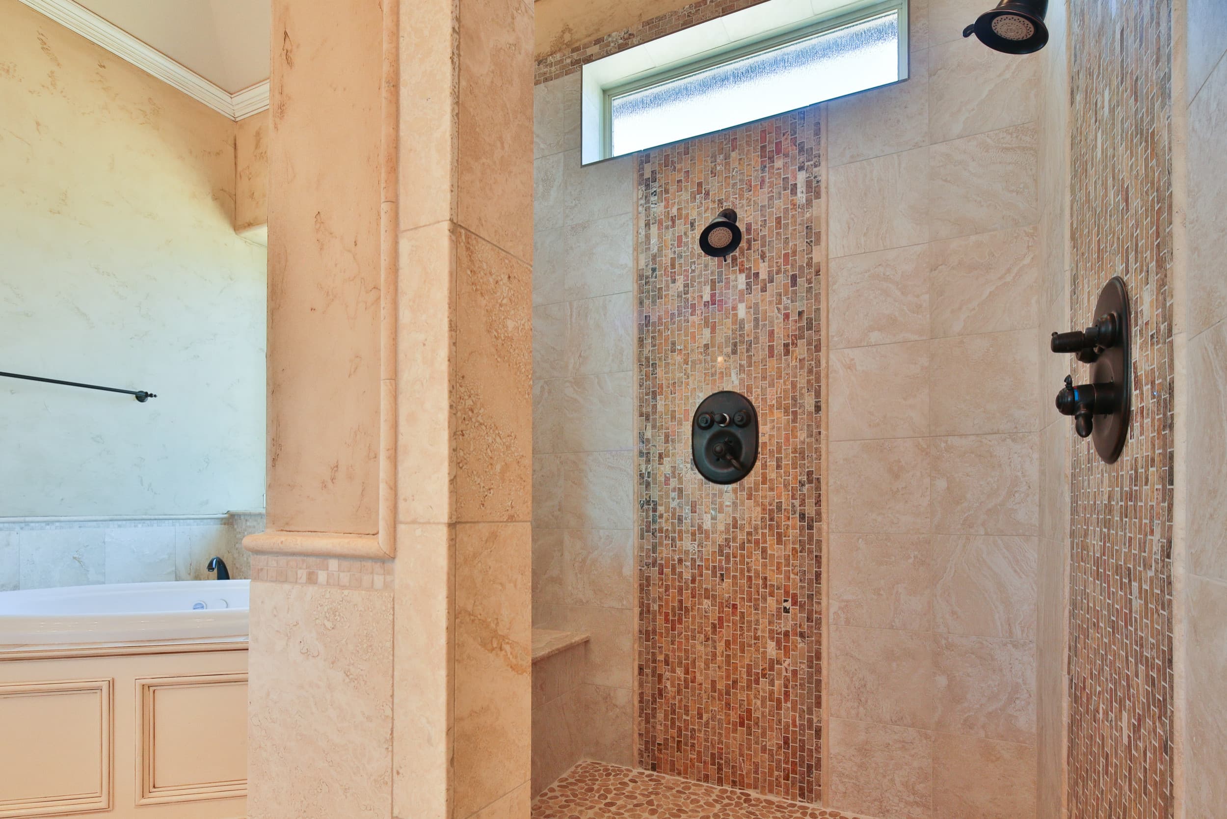 Walk-in shower with cream stone tile, vertical mosaic accent bands, dual bronze showerheads, pebble floor, corner bench, and high frosted window