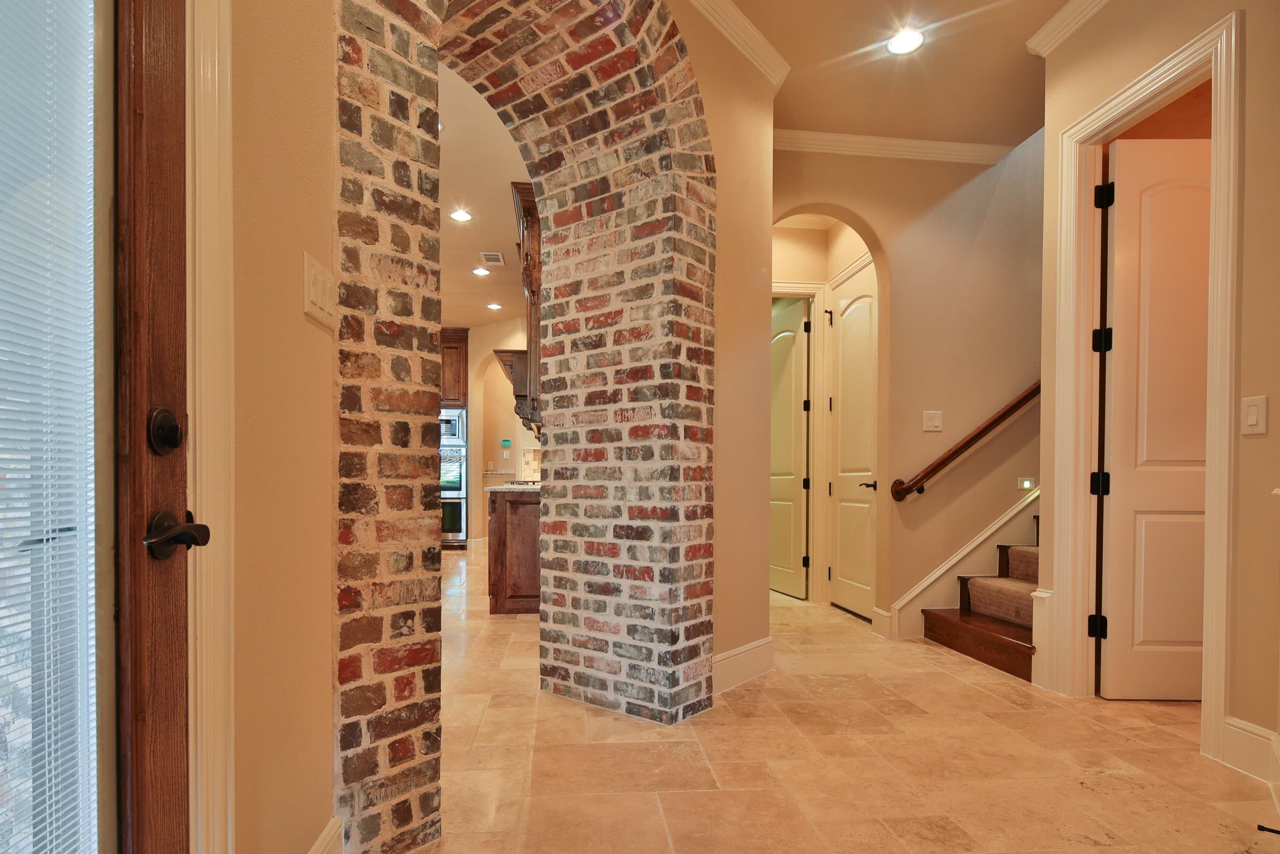 Travertine-floored hall with a brick archway into the kitchen, a carpeted staircase with wood handrail, and white paneled doors