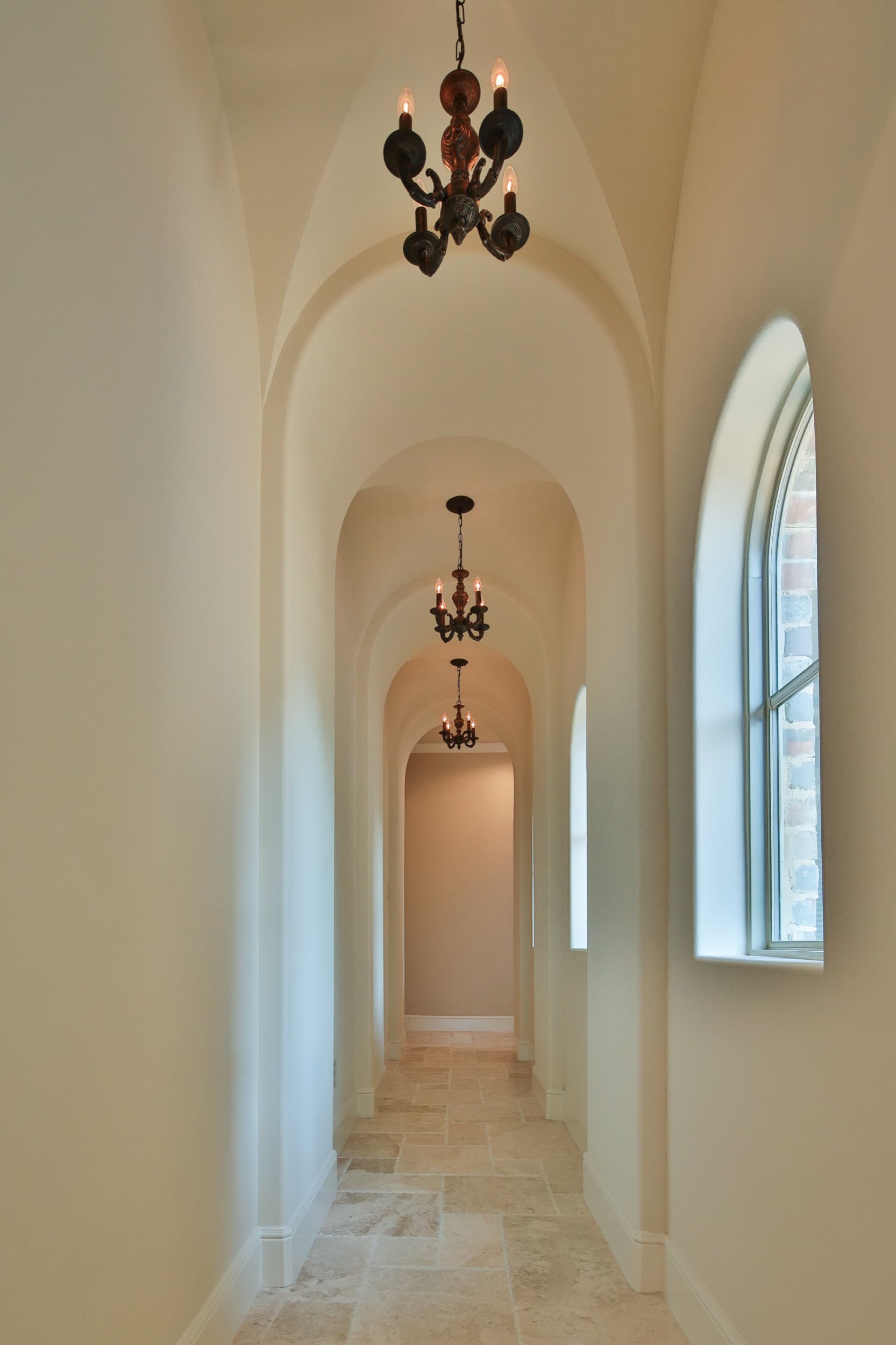Narrow groin-vaulted hallway with a series of arches, three bronze candle chandeliers, arched windows, and a travertine tile floor