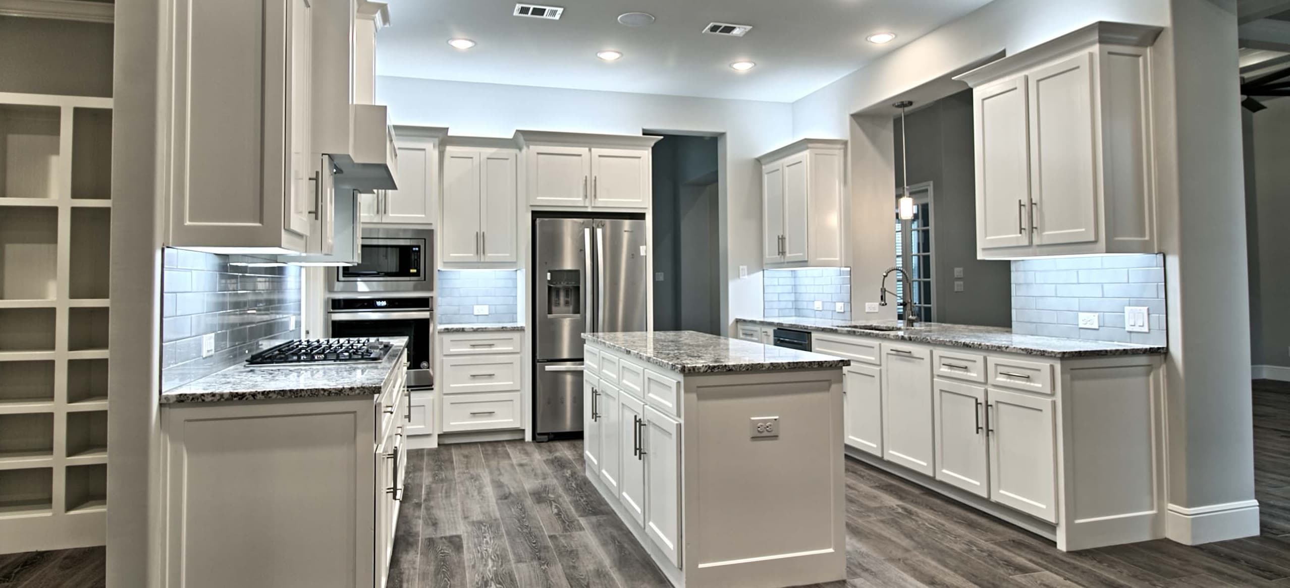 Wide kitchen view with white shaker cabinets, granite counters, glossy gray subway tile, stainless appliances, a center island and wood-look tile floor