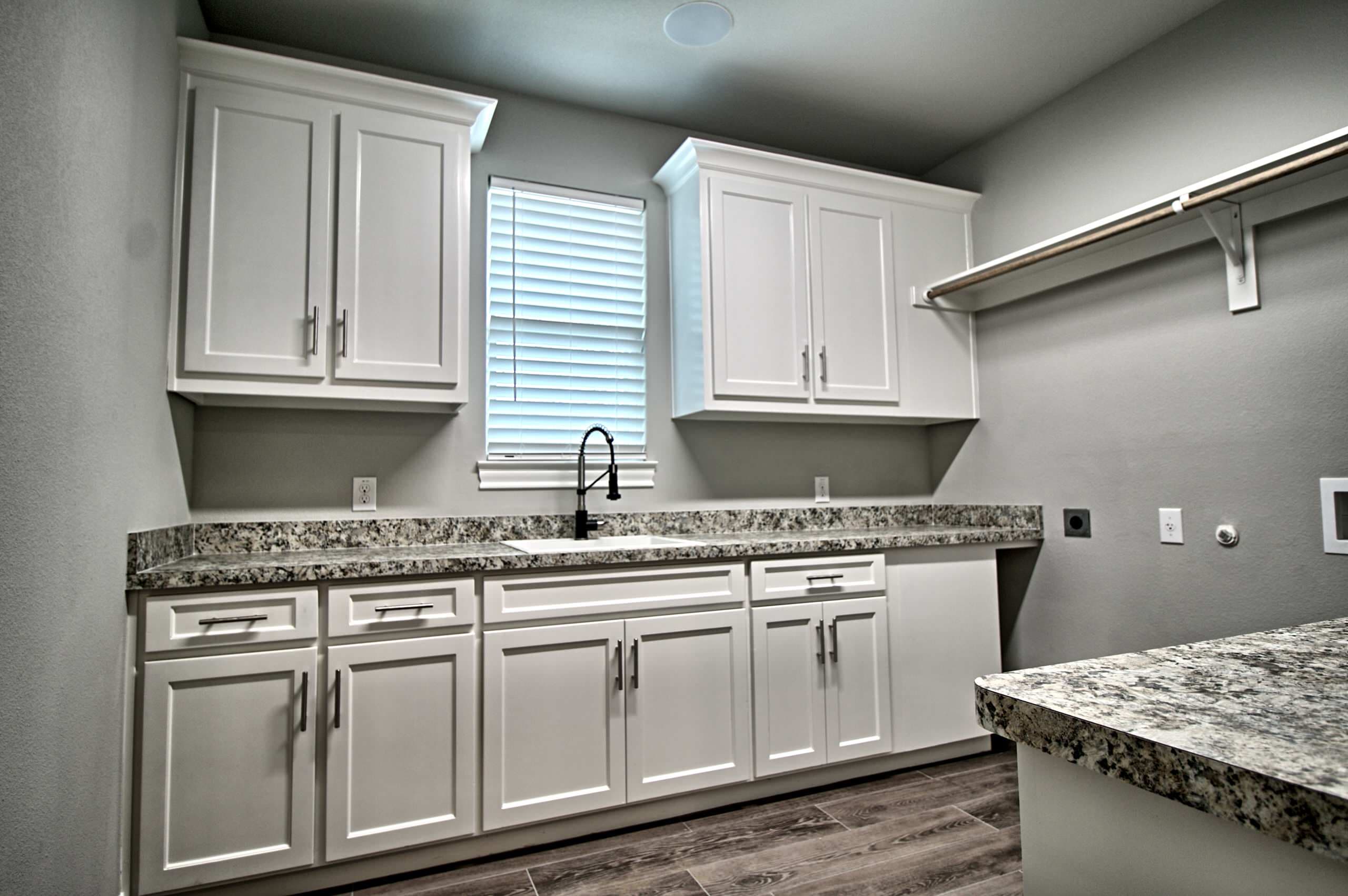 Laundry room with white shaker cabinets, granite counters, a white sink with black coil faucet, hanging rod and wood-look tile floor