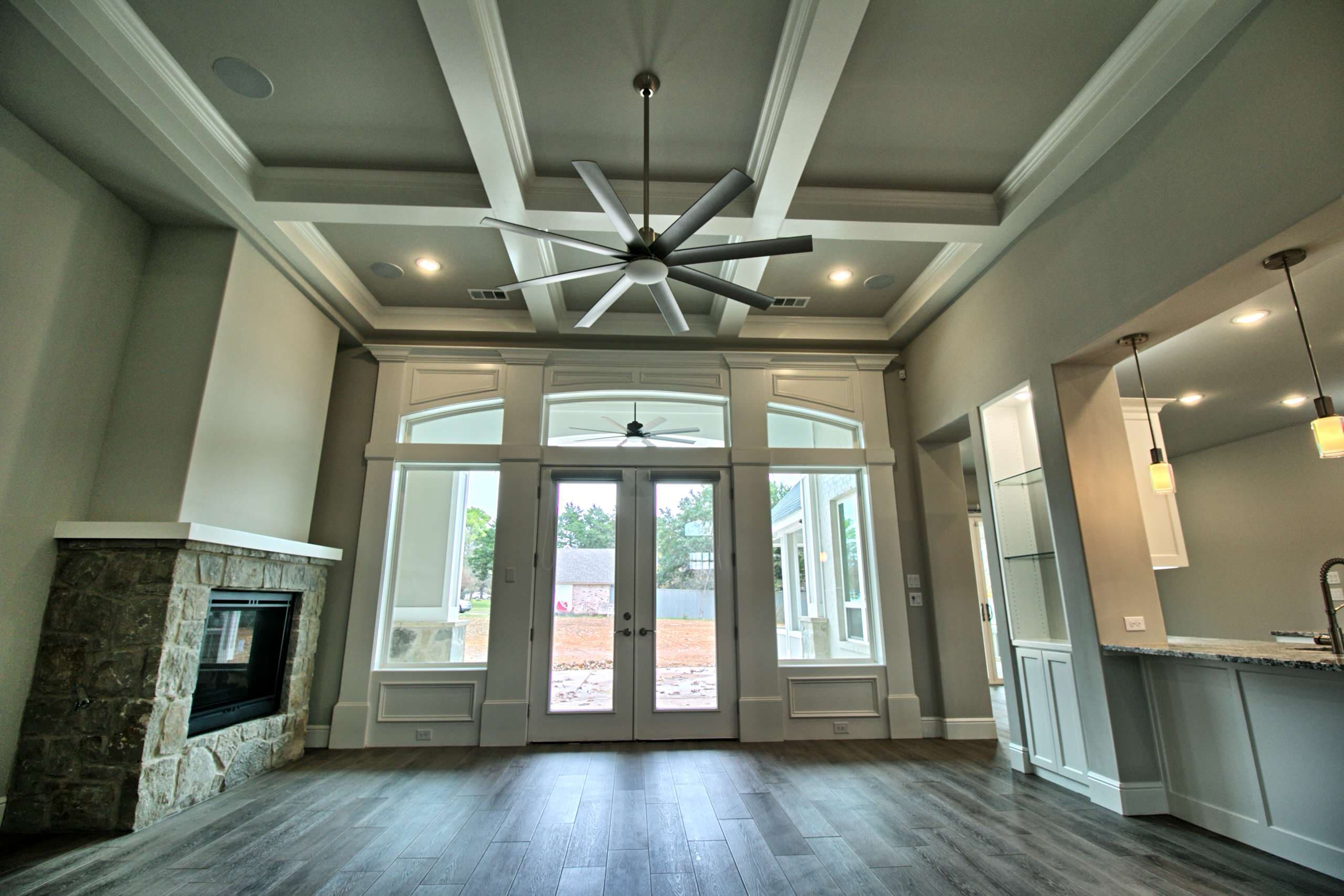 Living room with coffered ceiling, eight-blade ceiling fan, corner stone fireplace and French doors flanked by arched-transom windows in white paneled millwork