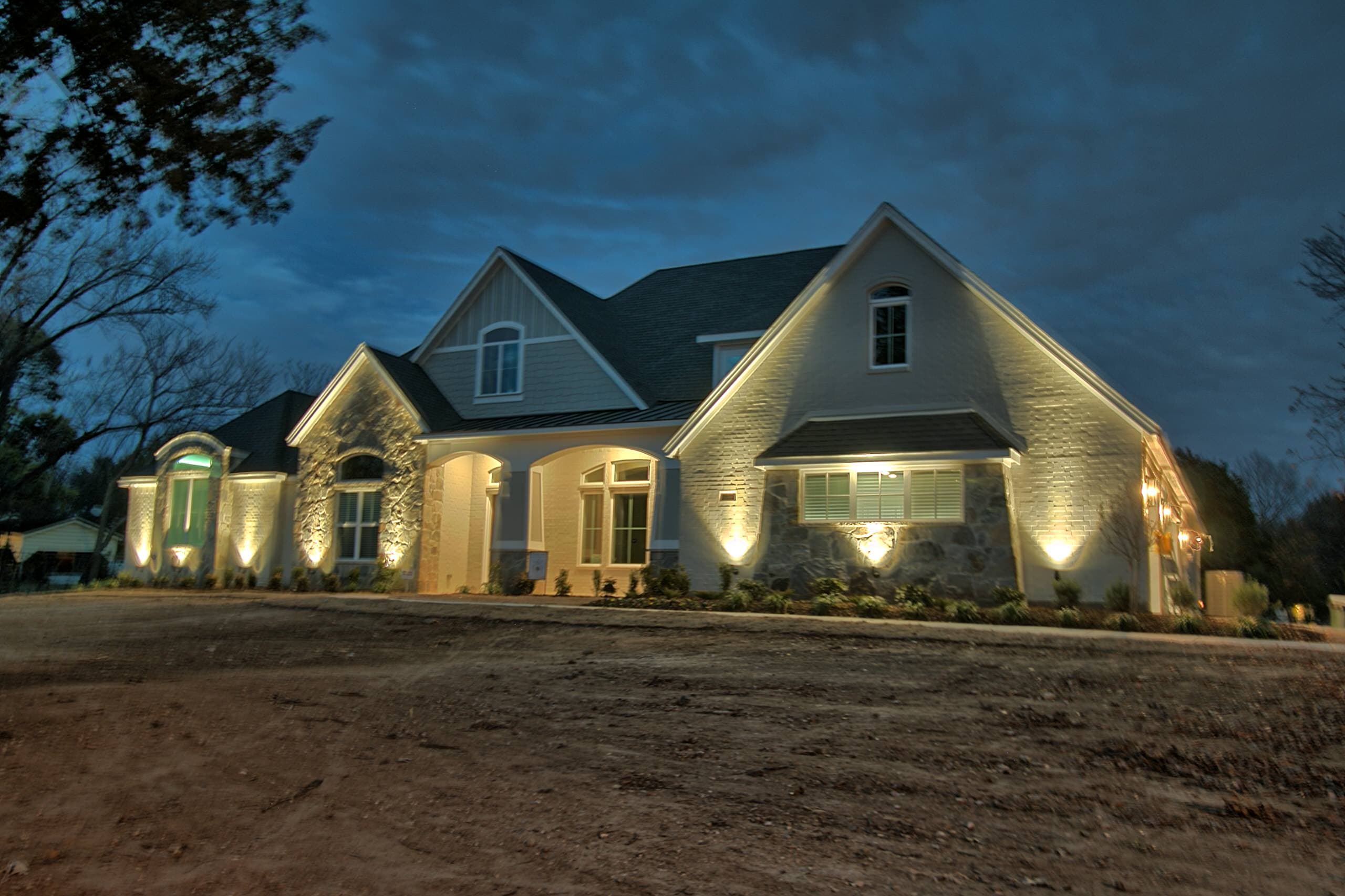 Front elevation at dusk with uplit painted brick and stone, gables with shake and board-and-batten siding, arched entry porch and foundation plantings