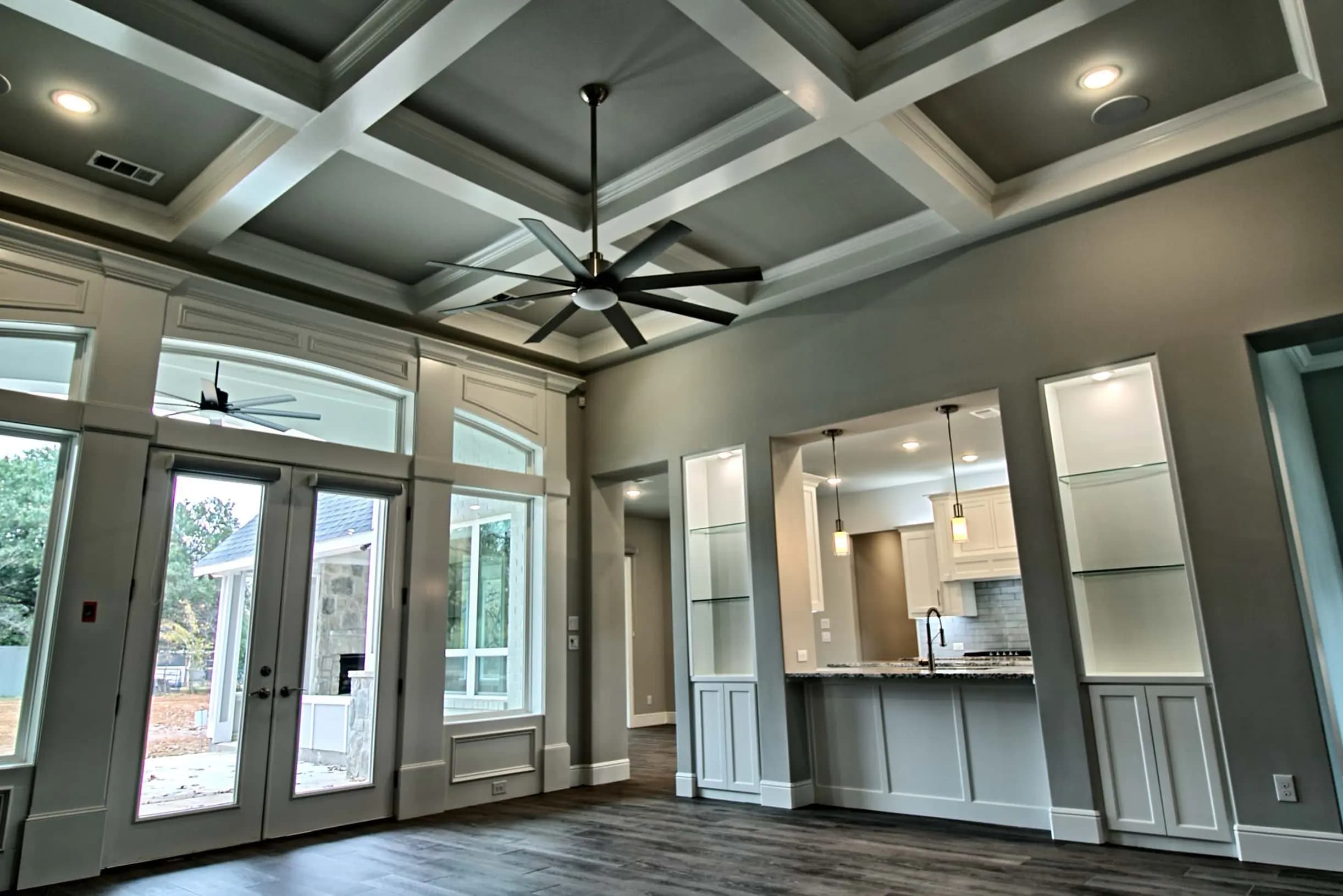 Modern open-concept living room with coffered ceiling, oversized ceiling fan, glass built-ins, and French doors leading to a stone patio. Hardwood flooring and pendant lighting accent the luxury Craftsman-style interior.