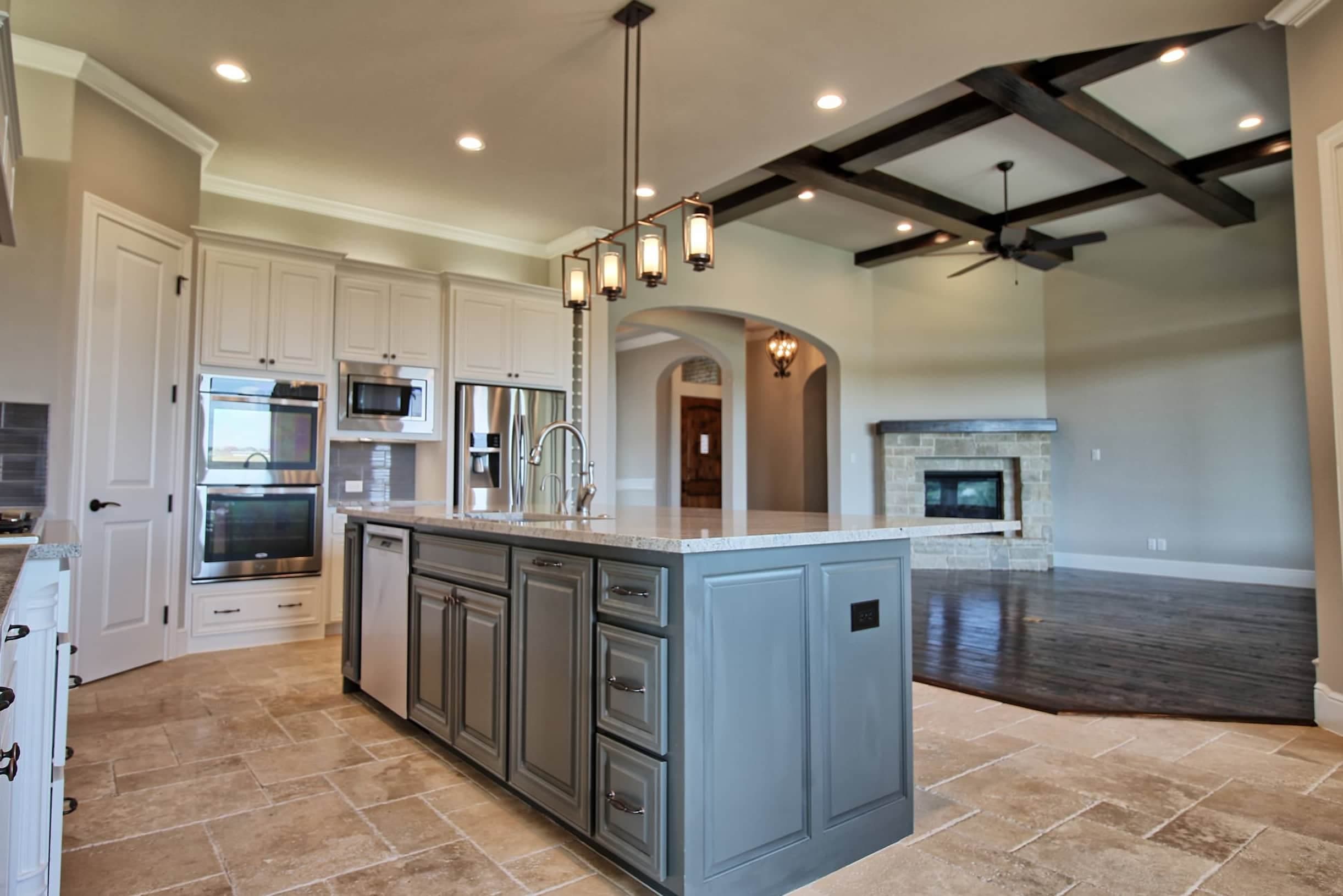 Modern custom kitchen by Oakmark Custom Homes with contrasting cabinetry, oversized island, pendant lighting, and open living room with stone fireplace.