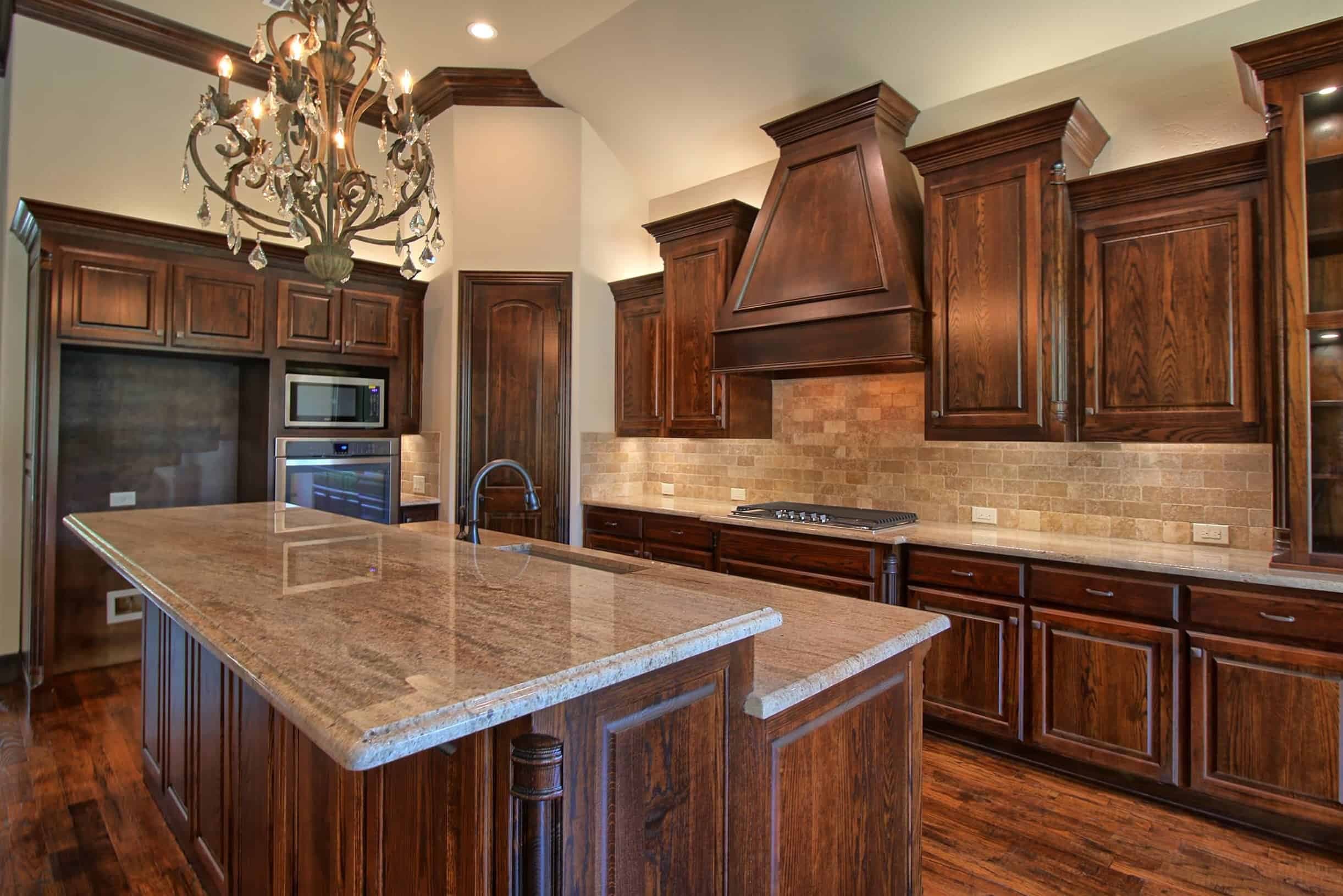 Elegant Oakmark Custom Homes kitchen with rich wood cabinetry, ornate chandelier, stone backsplash, and large granite island centerpiece.