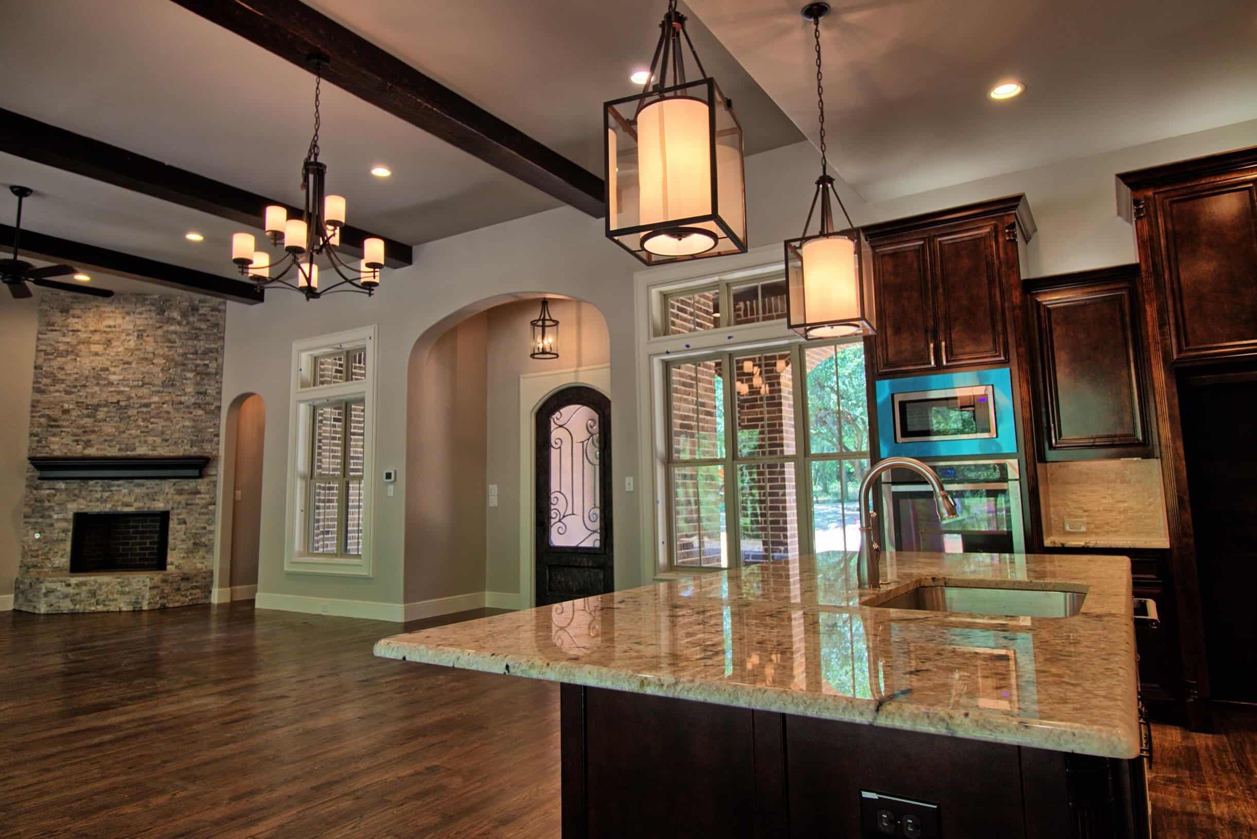 Luxury Oakmark Custom Homes living room and kitchen with stone fireplace, exposed beams, and granite island under designer pendant lights.