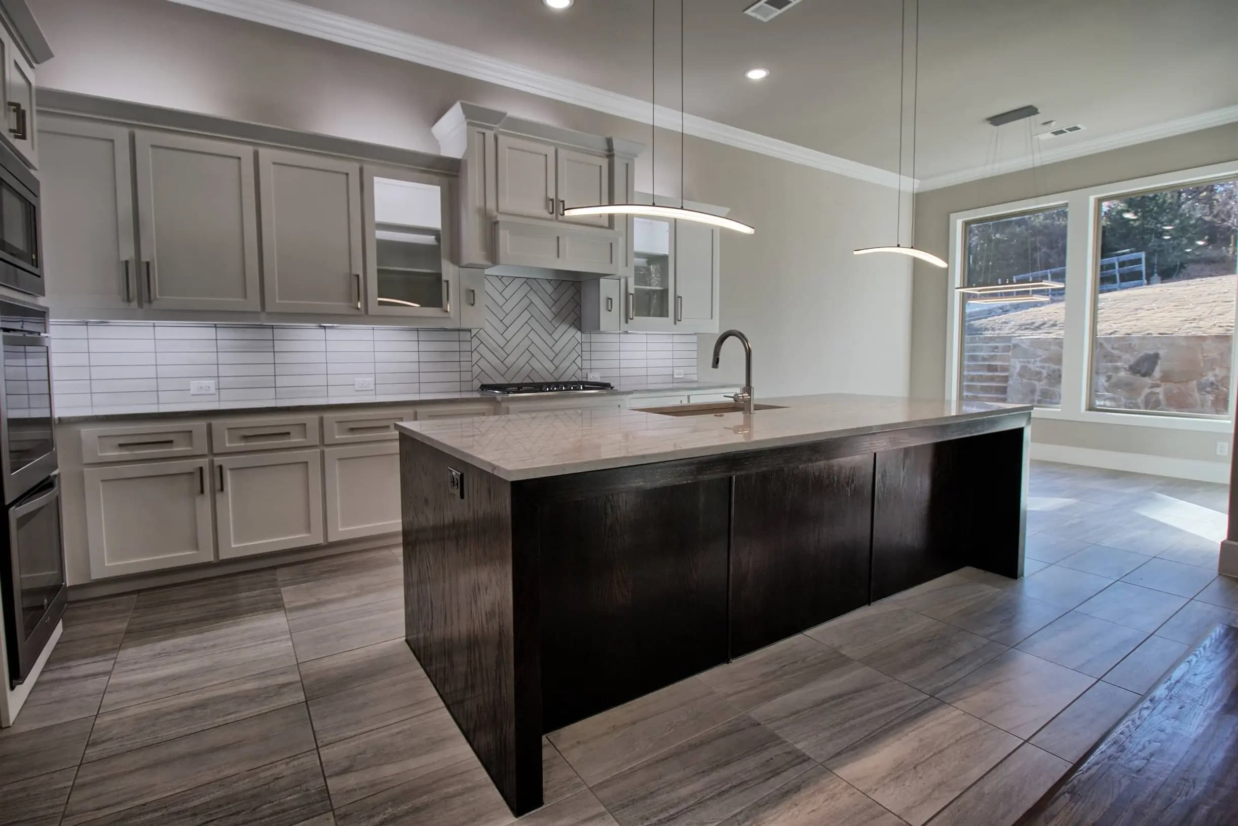 Contemporary kitchen with light gray shaker cabinets, herringbone tile backsplash, large picture windows, and a dark wood island with quartz countertop and built-in sink. Features modern pendant lighting and large format tile flooring.