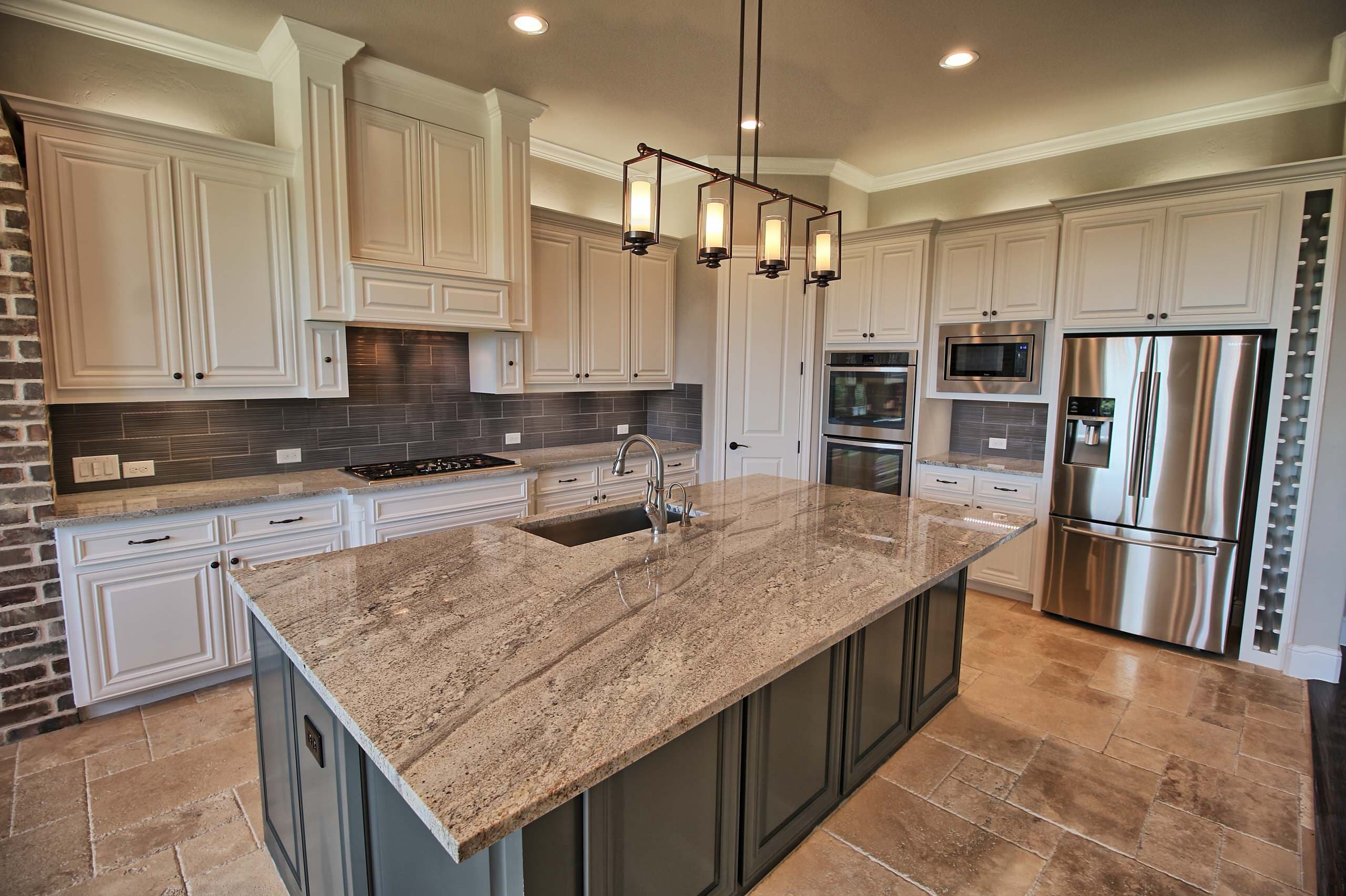 Kitchen with white cabinets, gray island with veined granite top, linear lantern pendant, stainless appliances, built-in wine rack, and travertine floors