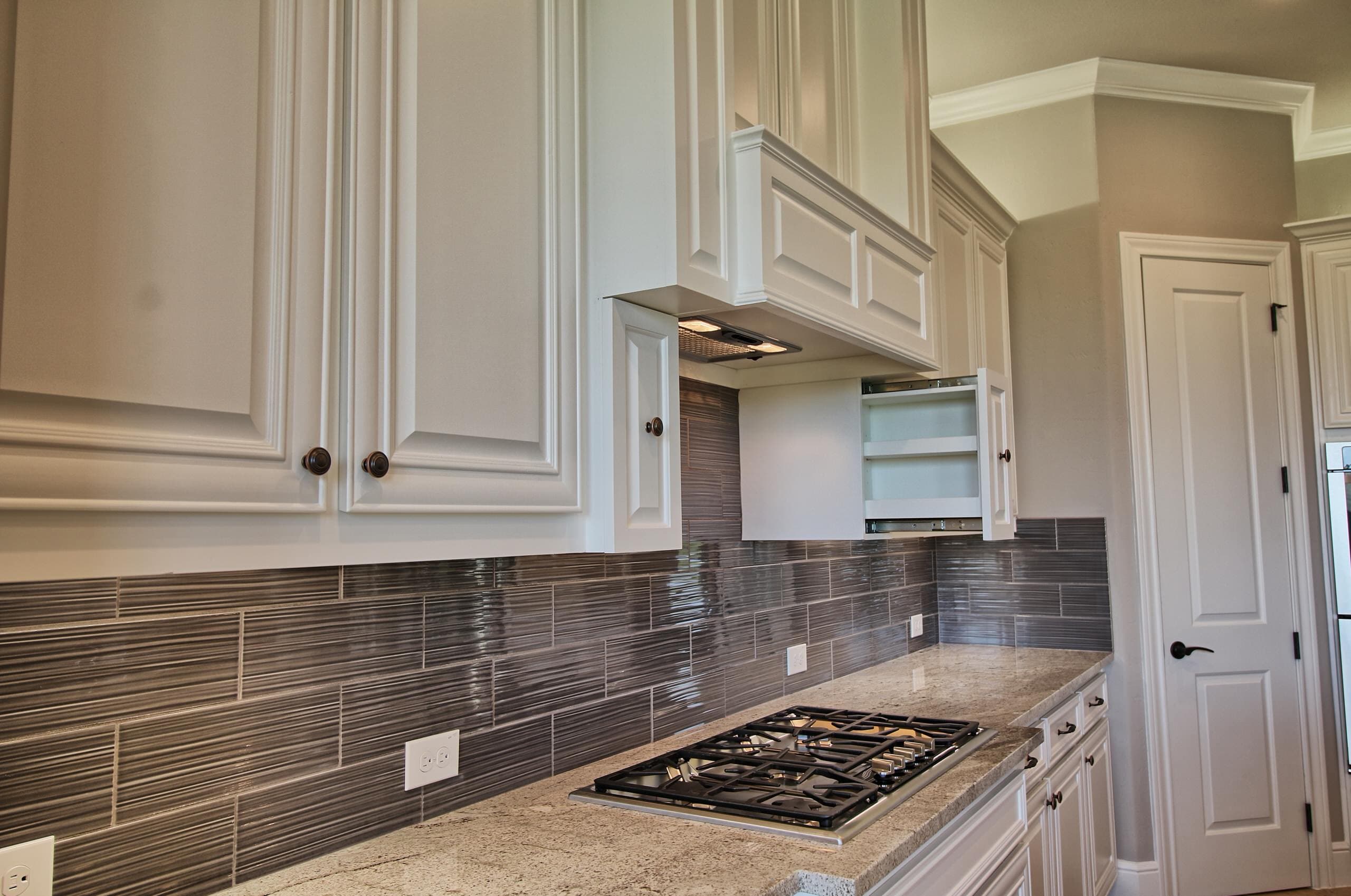 Gas cooktop in light granite beneath a cabinet hood, with textured gray plank tile backsplash, white raised-panel cabinets, and pull-out spice rack