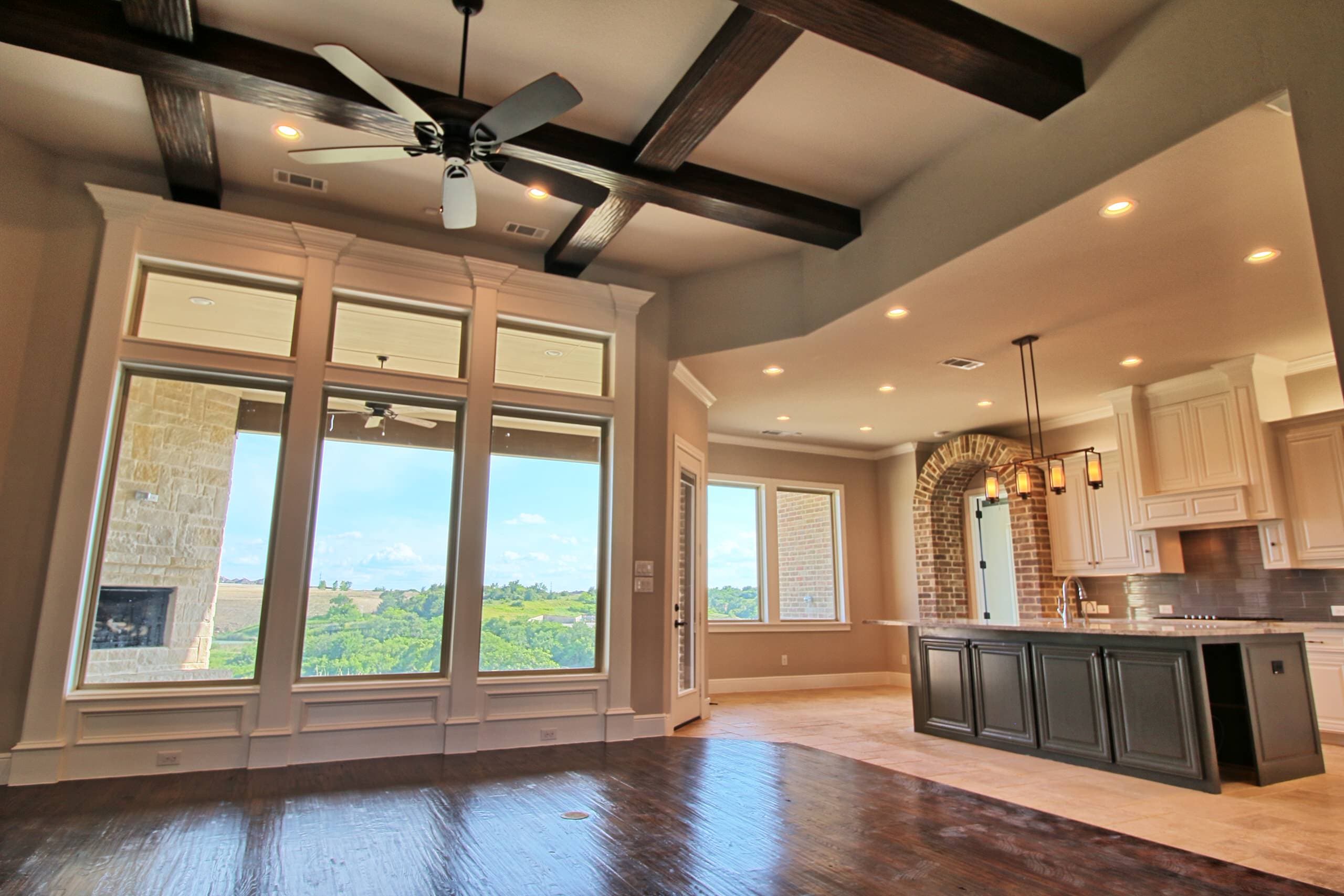 Great room with dark wood ceiling beams and transom-topped windows facing a covered patio with stone fireplace, open to the kitchen