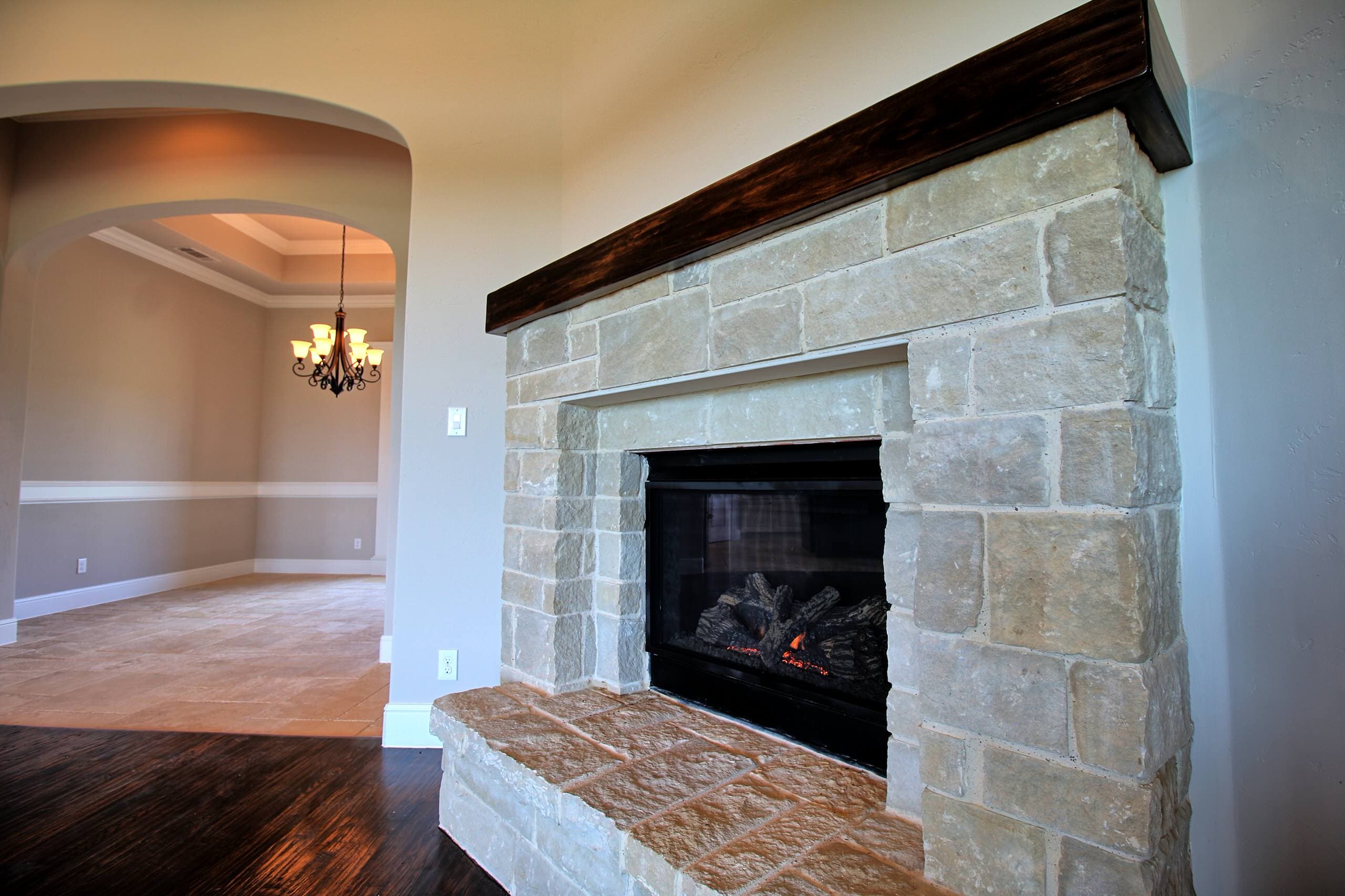 Cream cut-stone fireplace with raised hearth, dark wood beam mantel, and glass-front firebox, beside an arched opening to a dining room