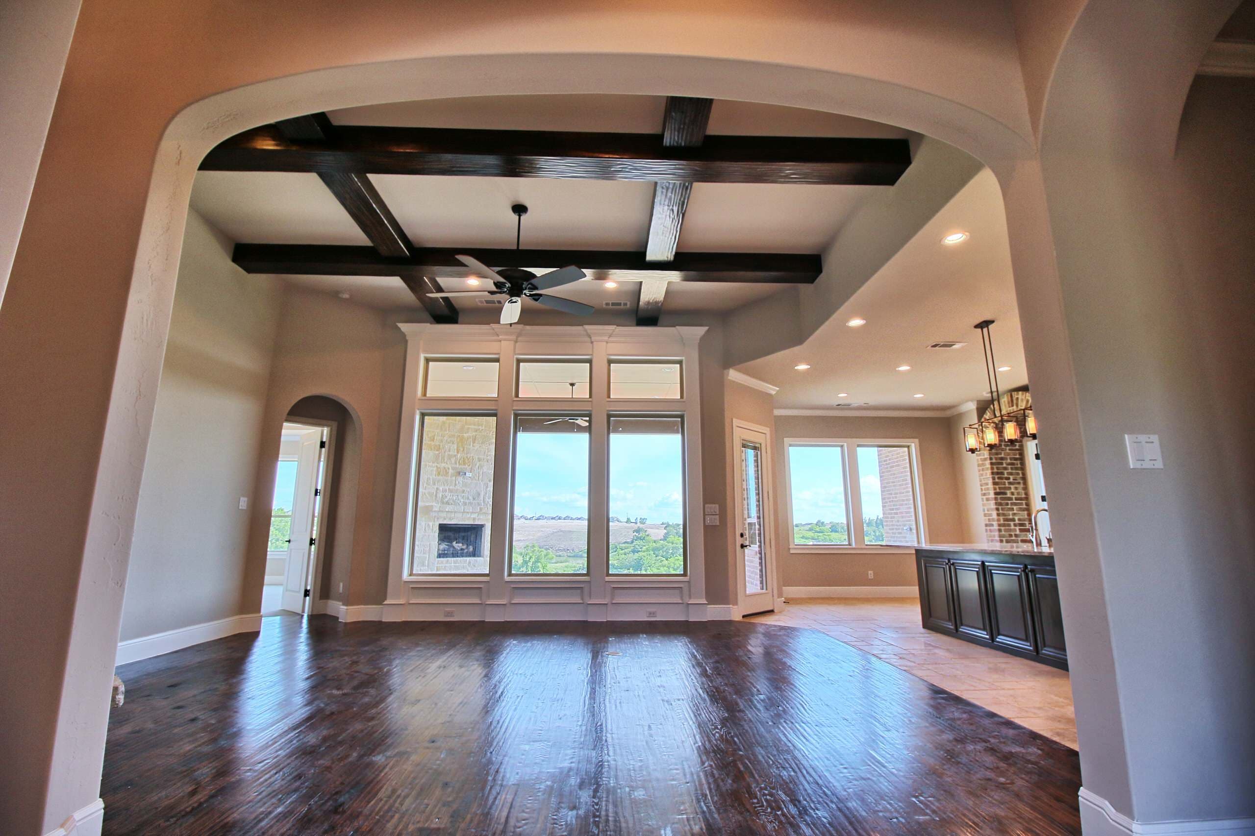 Great room framed by an arch, with dark wood ceiling beams, hand-scraped hardwood, and tall windows facing a covered patio and hills