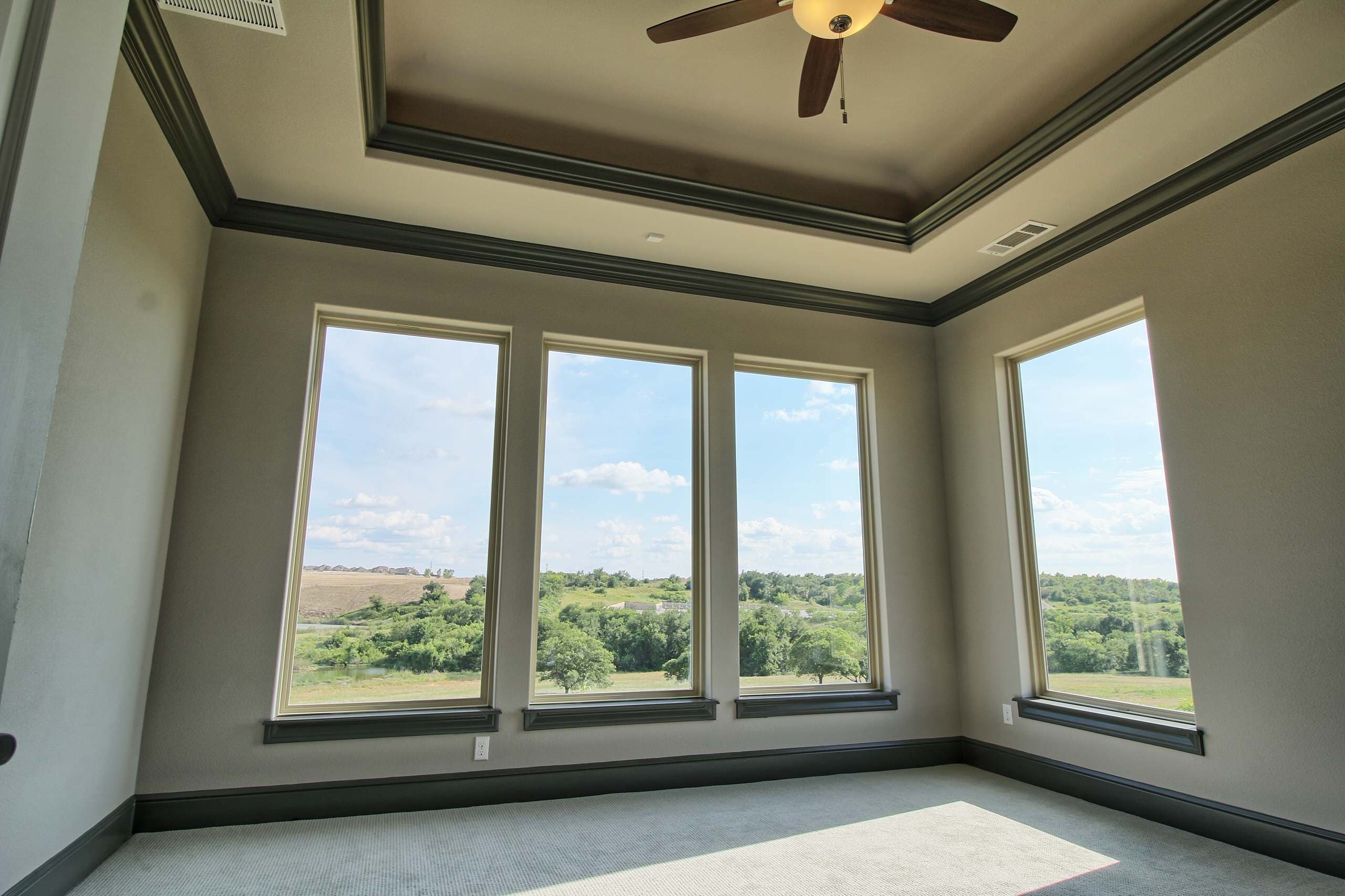 Carpeted bedroom with tray ceiling, charcoal crown molding and baseboards, ceiling fan, and four tall windows overlooking wooded hills