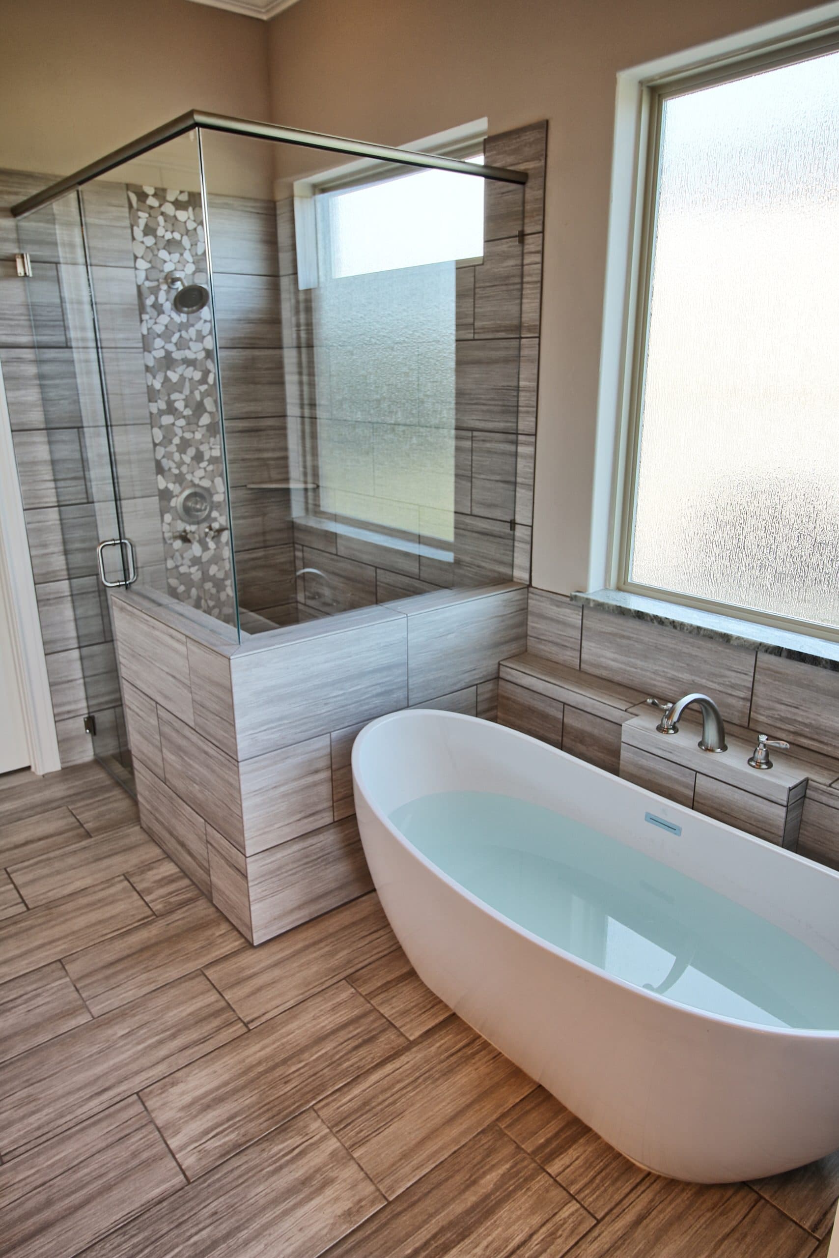 Freestanding soaking tub and frameless glass shower with striated plank tile, a river pebble accent strip, brushed nickel fixtures, and frosted windows