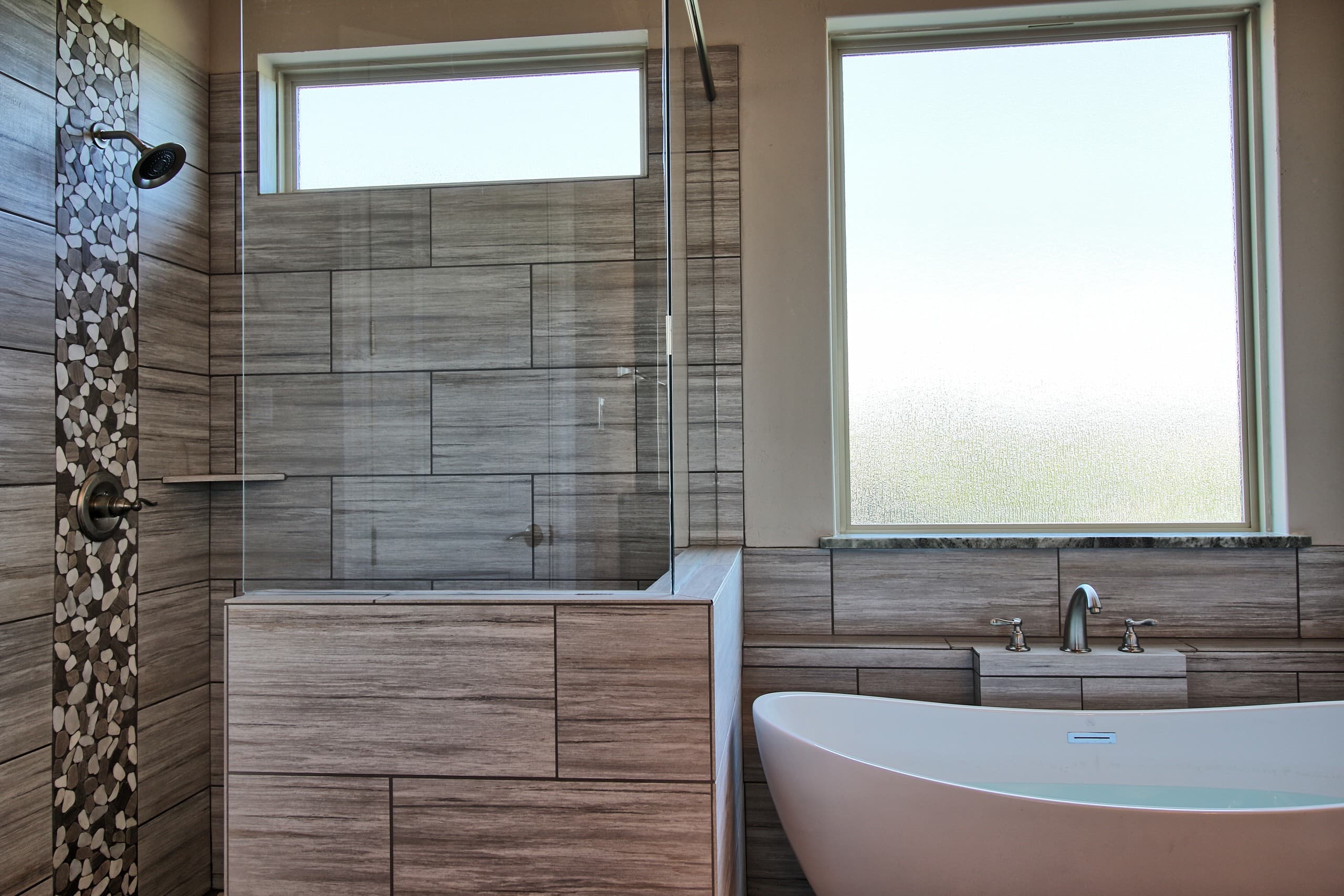 Shower with glass panel over a tiled half wall and river pebble accent strip, beside a freestanding tub under a frosted window