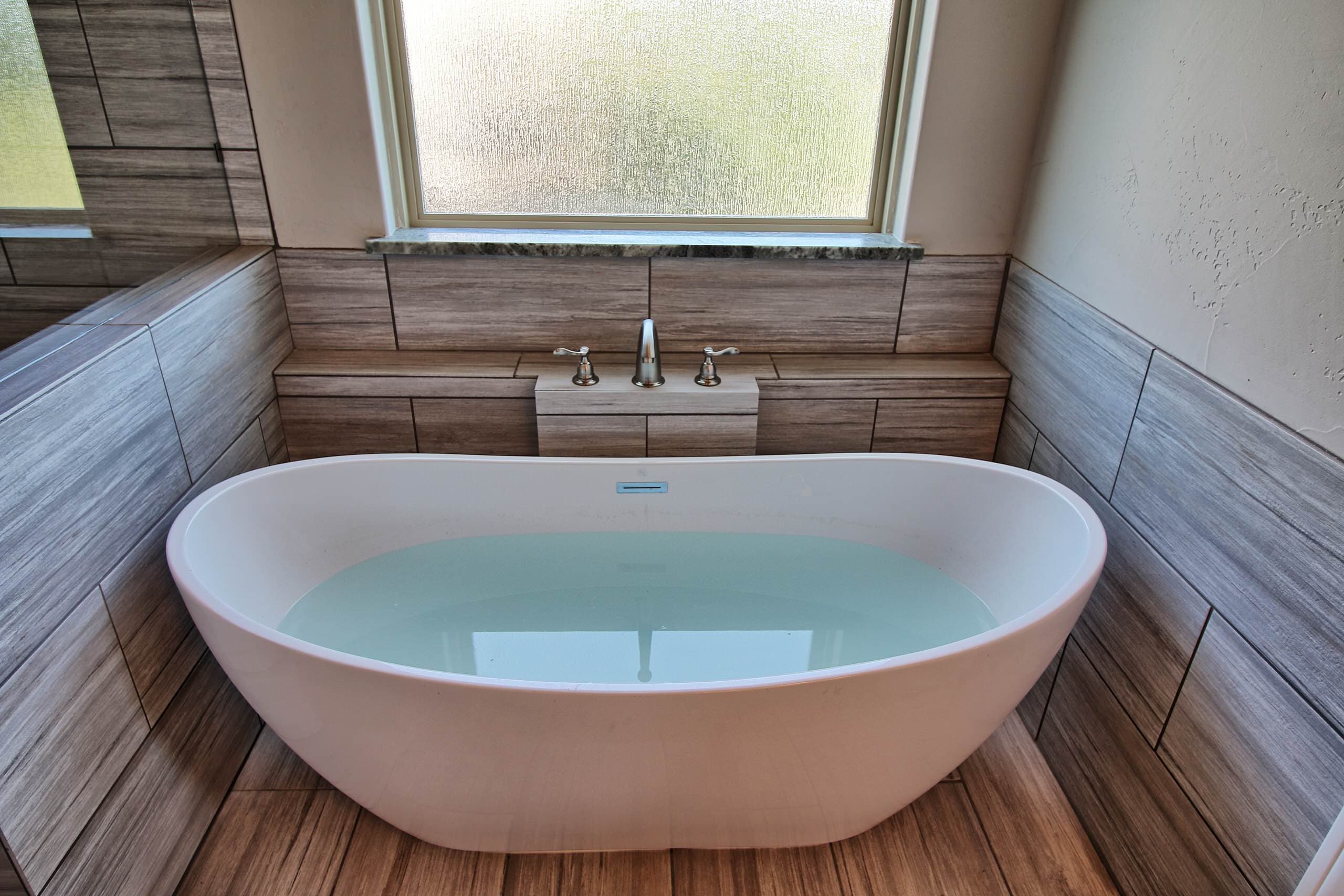 Freestanding oval soaking tub with brushed nickel deck-mount faucet on a tiled ledge, beneath a frosted window with stone sill