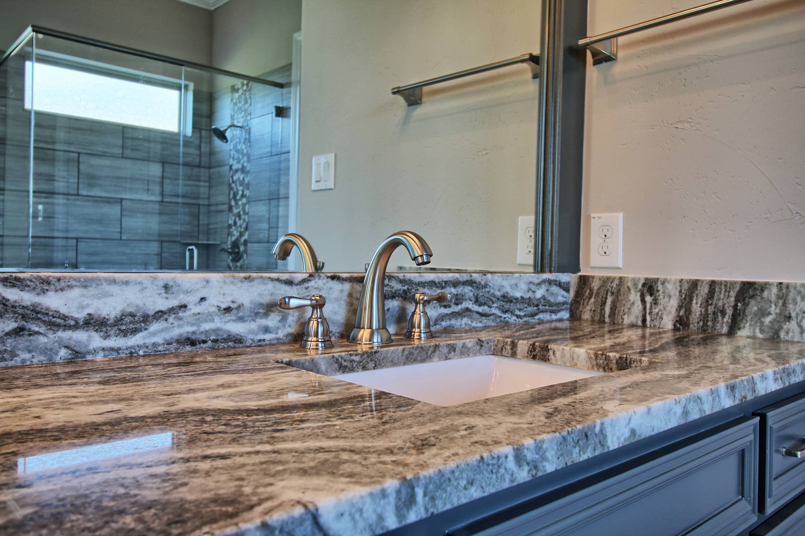 Vanity detail with swirled gray stone countertop and backsplash, undermount sink, brushed nickel widespread faucet, and charcoal cabinetry