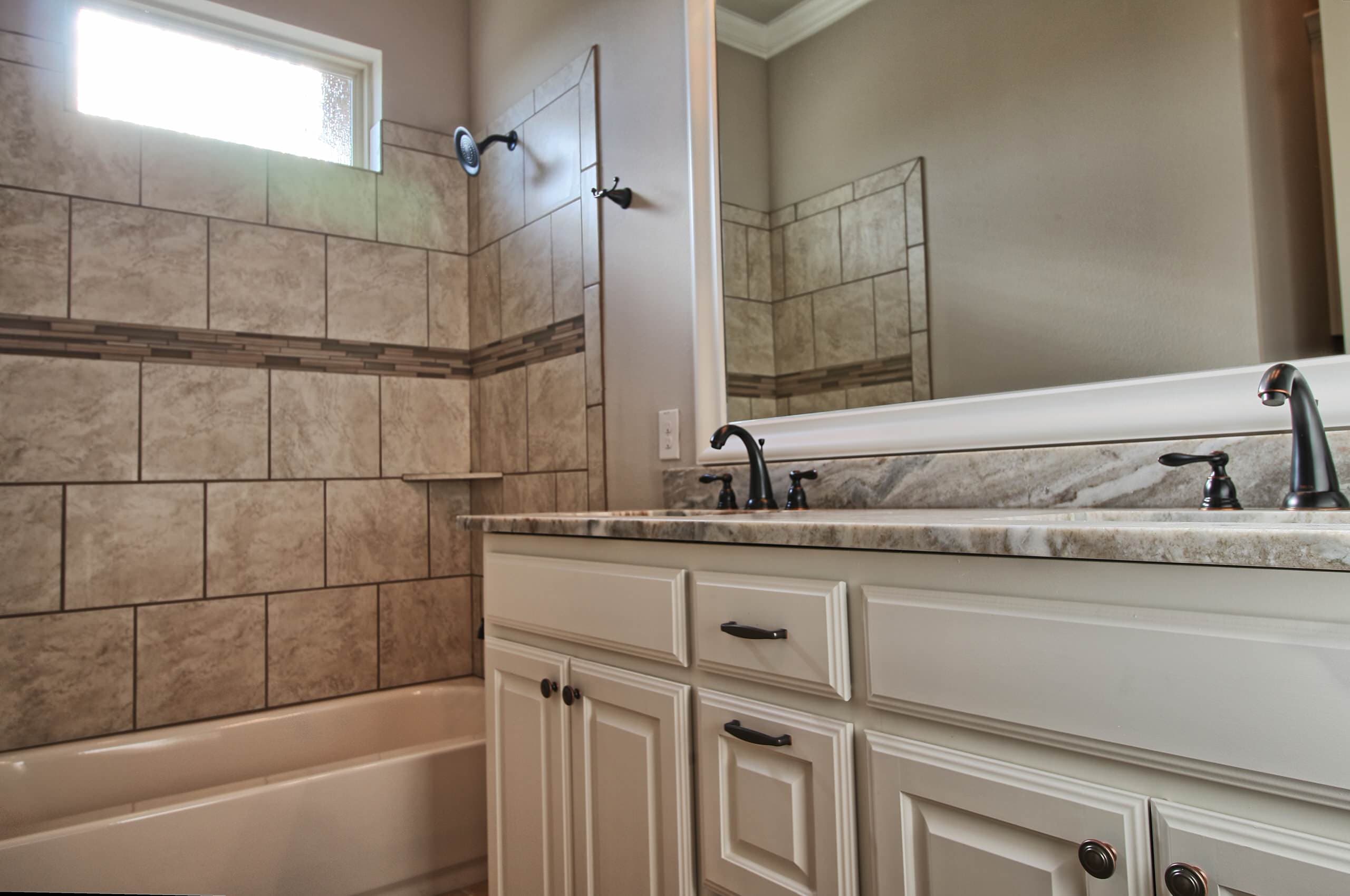 Bathroom with tub and shower surround of beige stone-look tile with mosaic band, white double vanity, veined stone counter, and bronze faucets