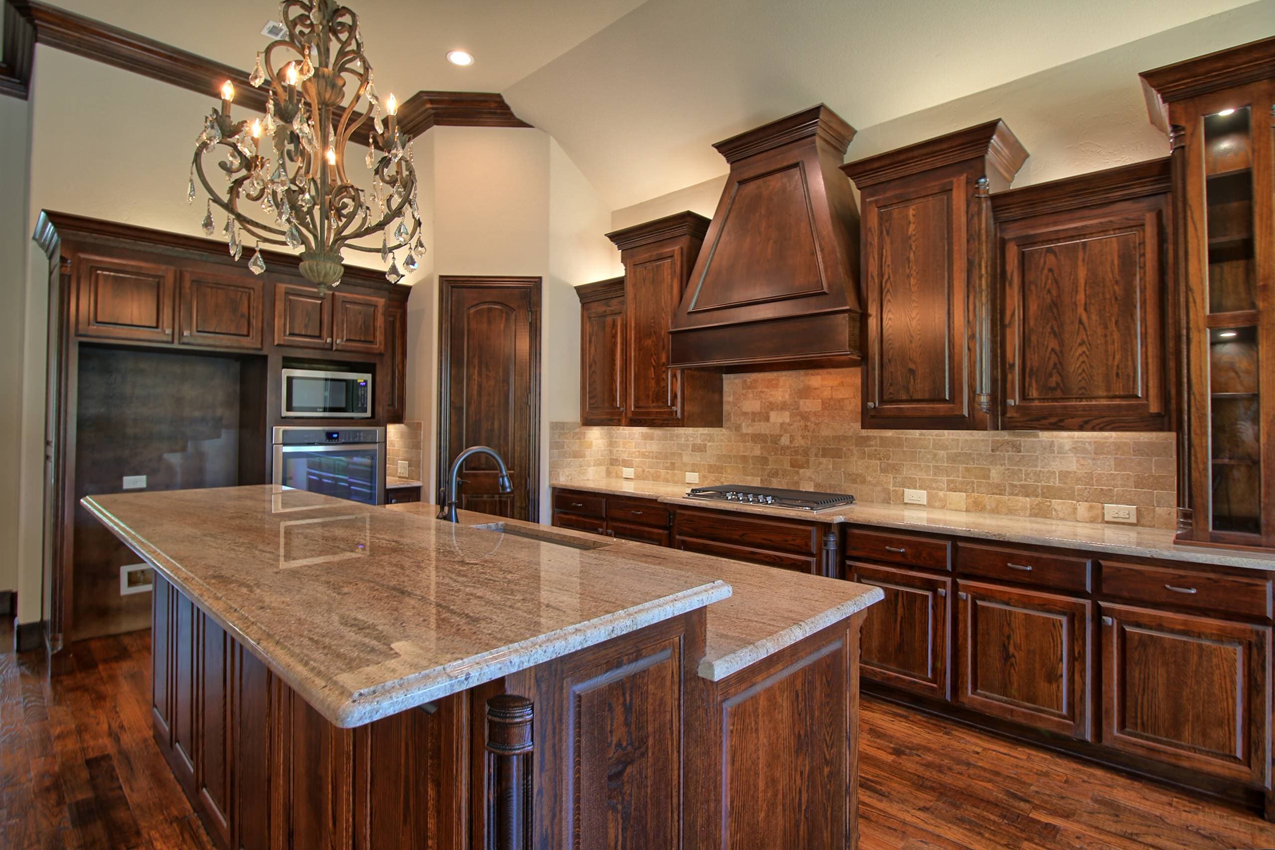 Kitchen with dark stained oak cabinetry, wood range hood, travertine subway backsplash, granite island with sink, crystal chandelier and hardwood floors
