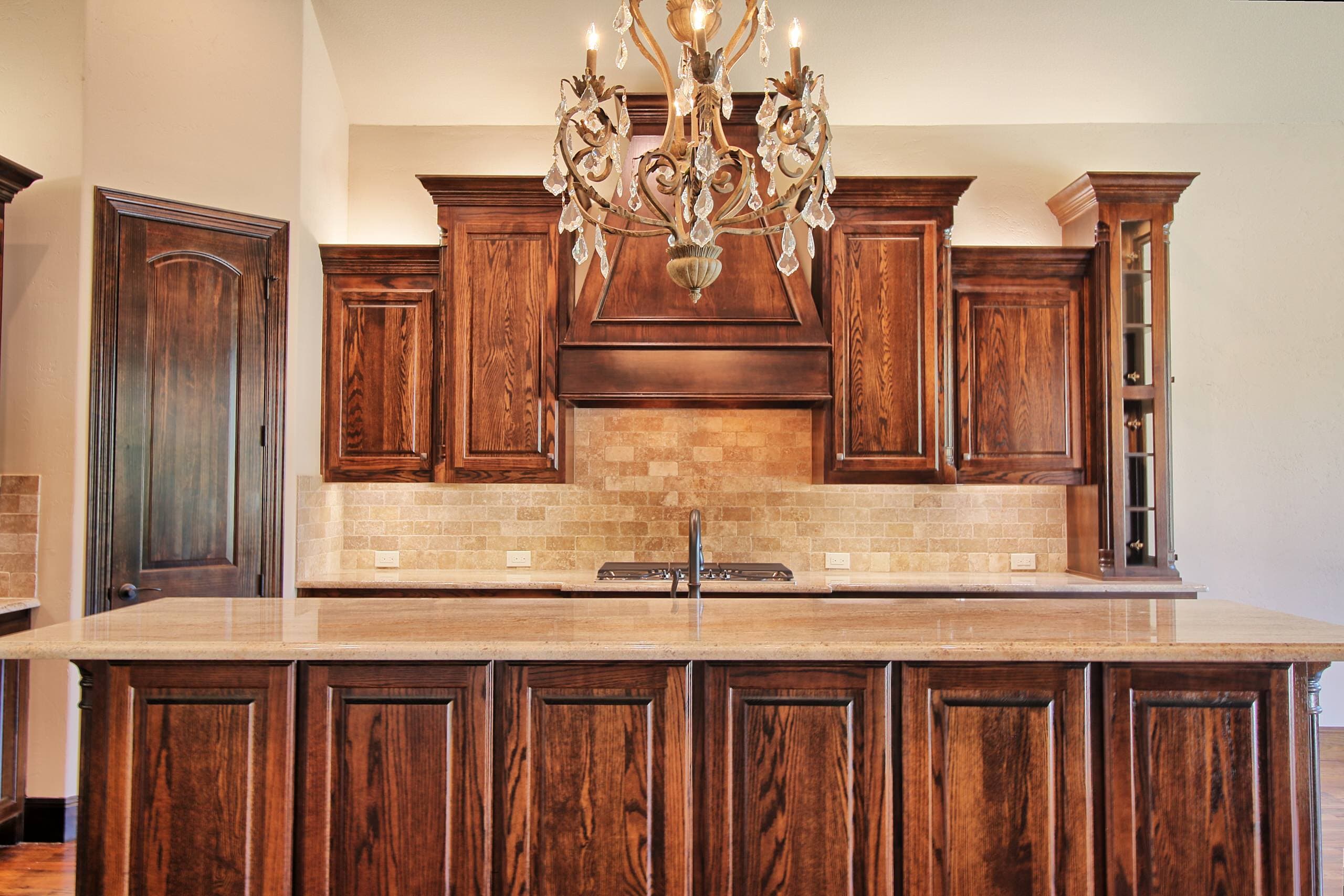 Kitchen island with paneled oak base and cream granite top under a crystal chandelier, facing an oak range hood and travertine backsplash