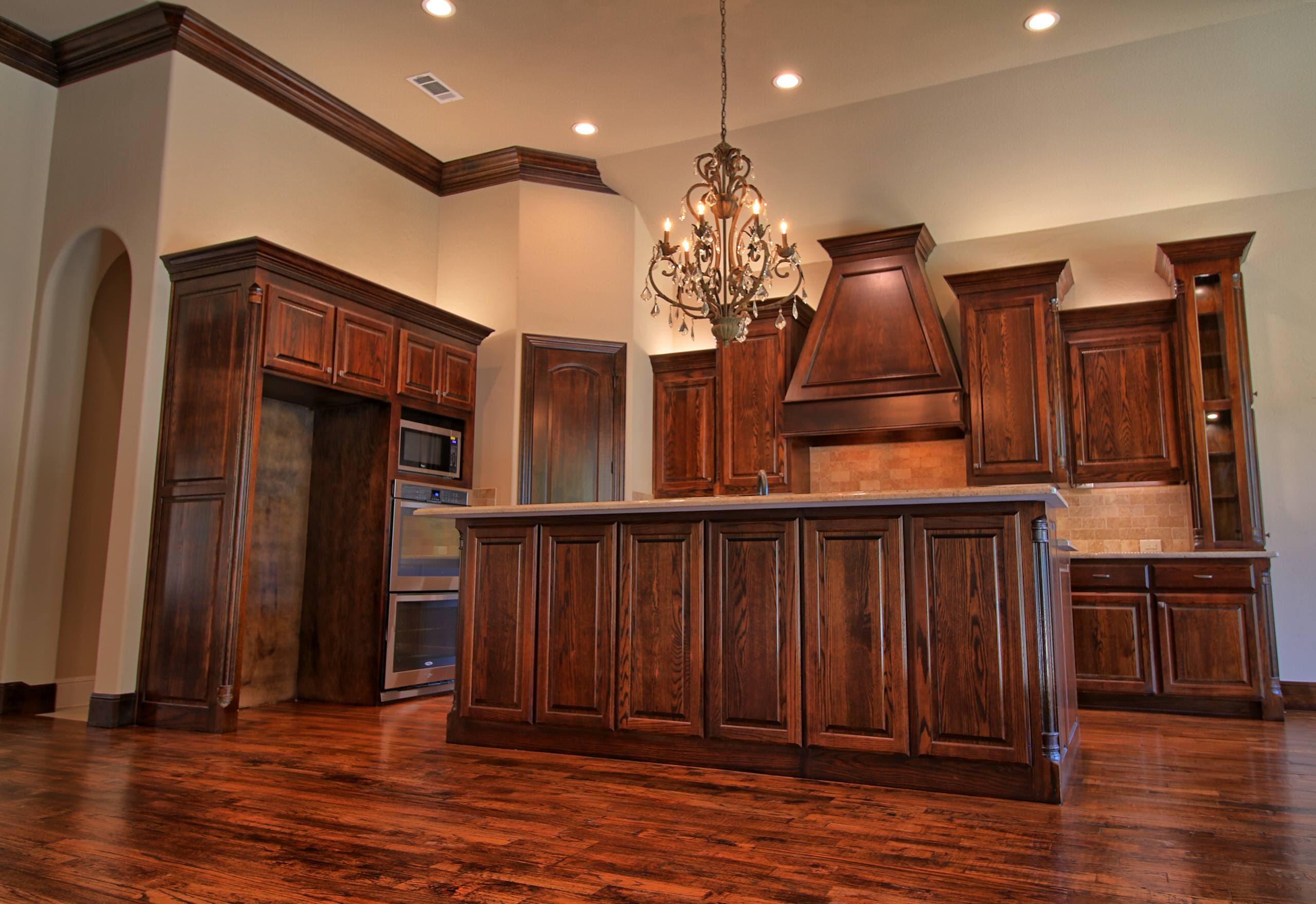 Kitchen with stained oak cabinetry and crown molding, paneled island, tapered wood hood, double wall ovens, crystal chandelier and hand-scraped hardwood floors
