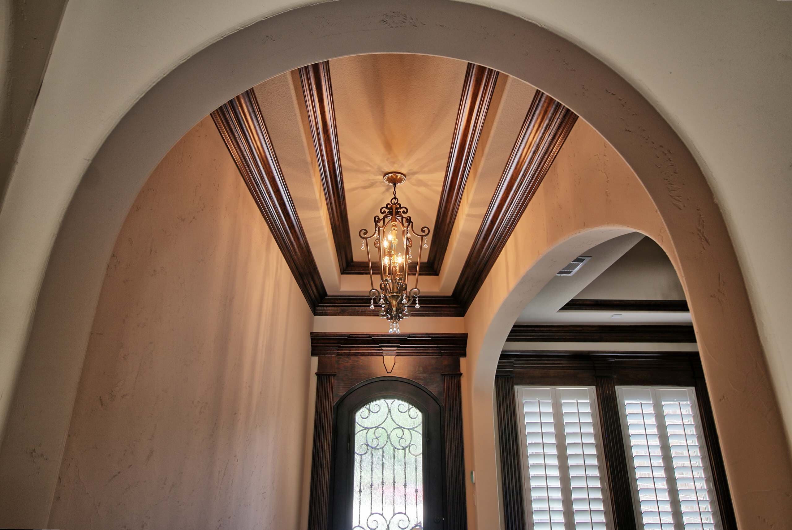 Entry viewed through a plaster arch, with tiered stained wood ceiling trim, iron lantern chandelier and arched front door with iron scrollwork