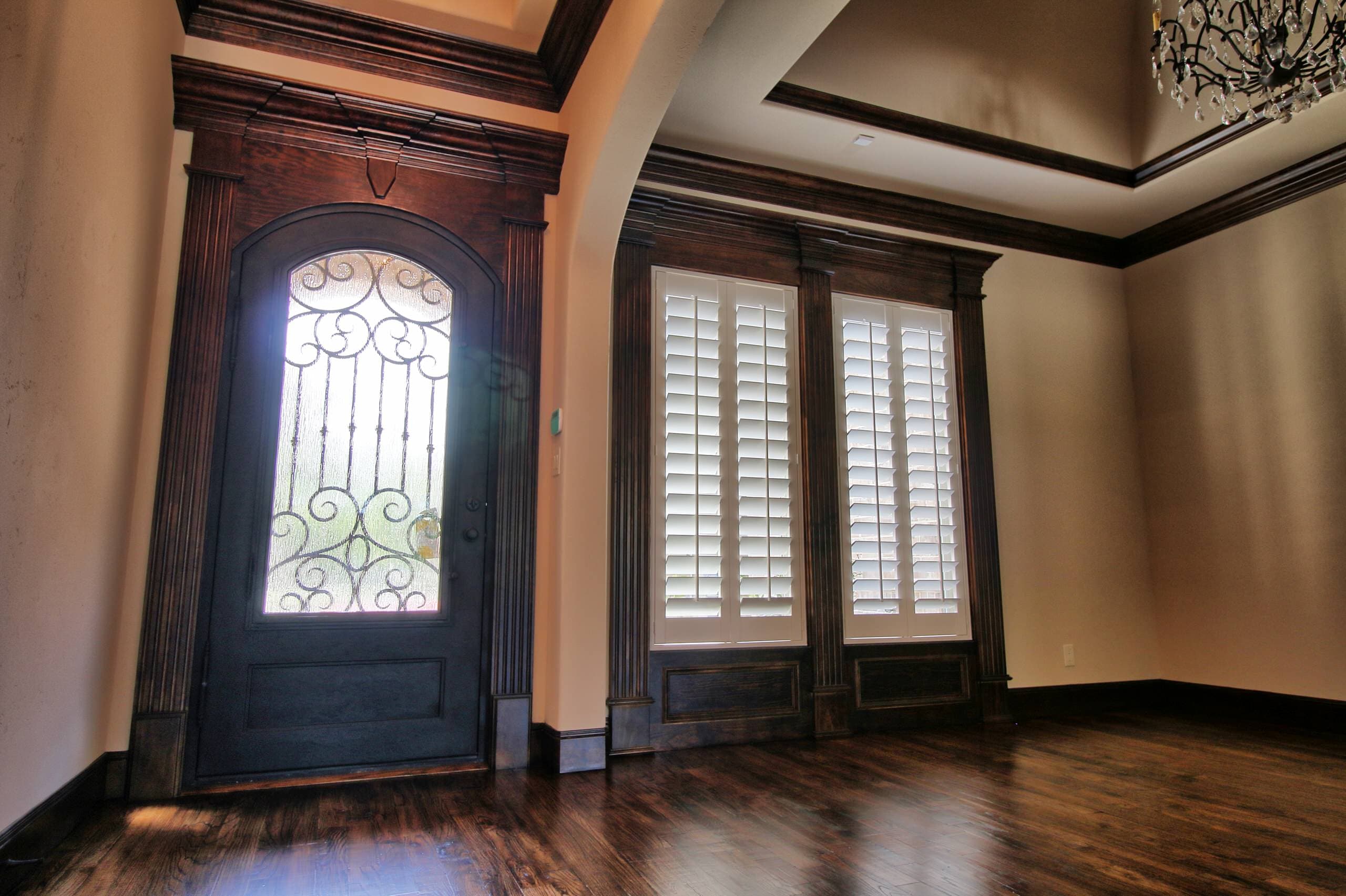 Foyer with arched iron and glass front door in stained wood casing, beside a room with shuttered windows and hand-scraped hardwood floors