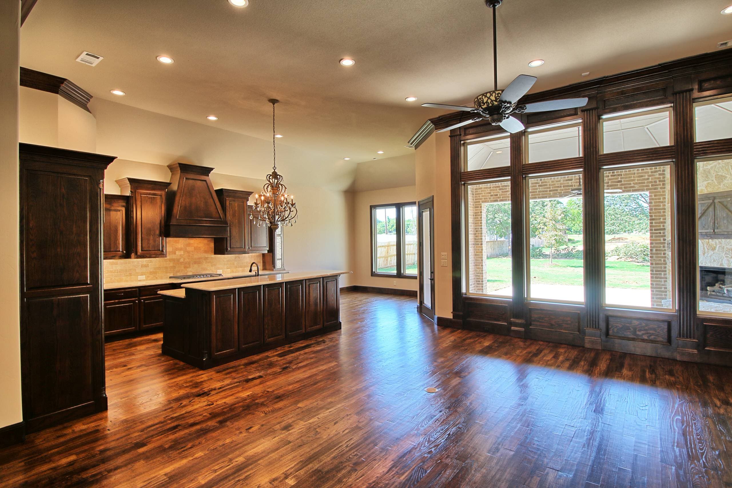 Great room with hand-scraped hardwood floors, ceiling fan, stained wood framed window wall facing a covered patio, and an open kitchen