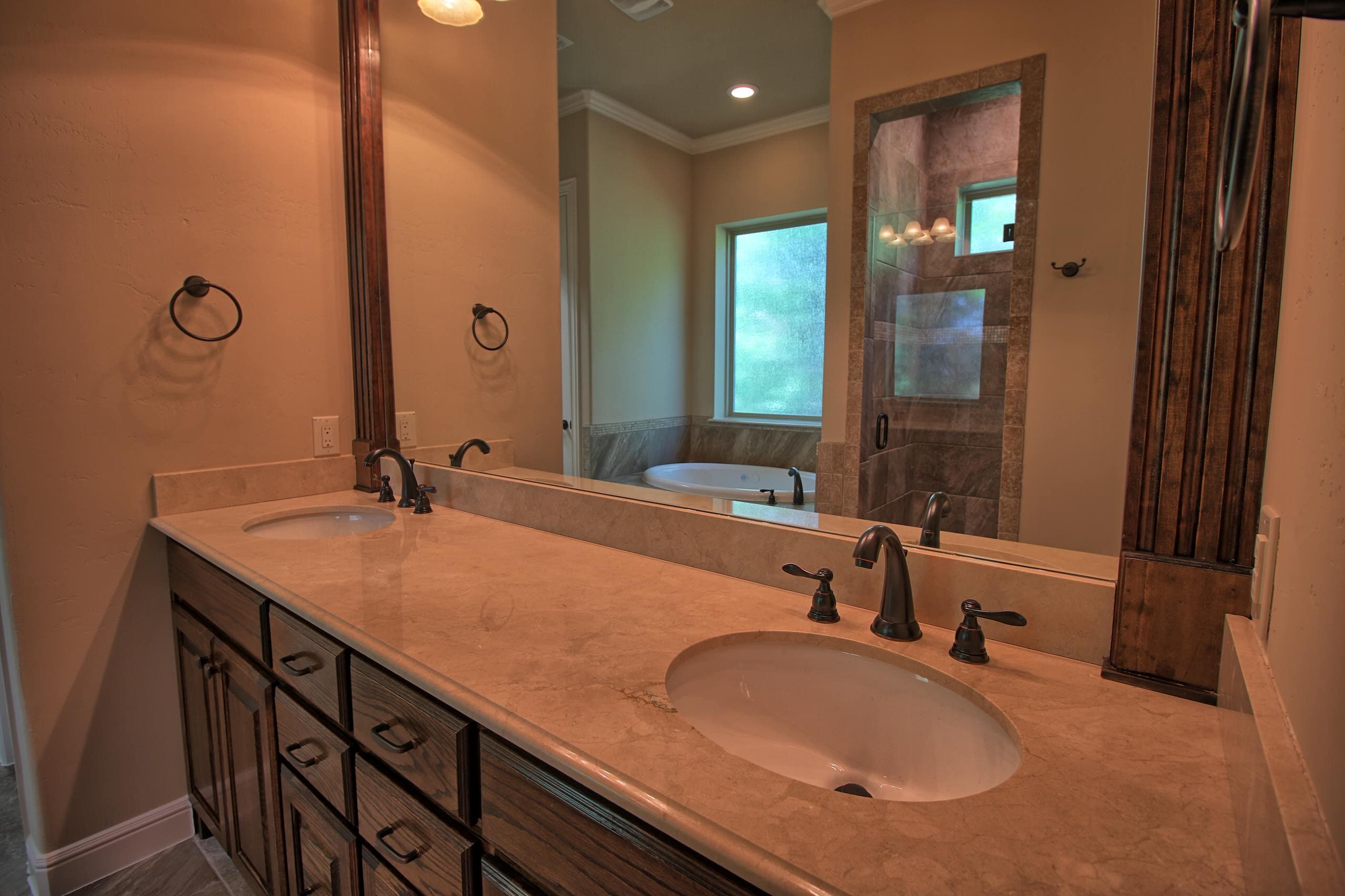 Double vanity with stained oak cabinets, cream marble top and bronze faucets, the mirror reflecting a jetted tub and tiled shower
