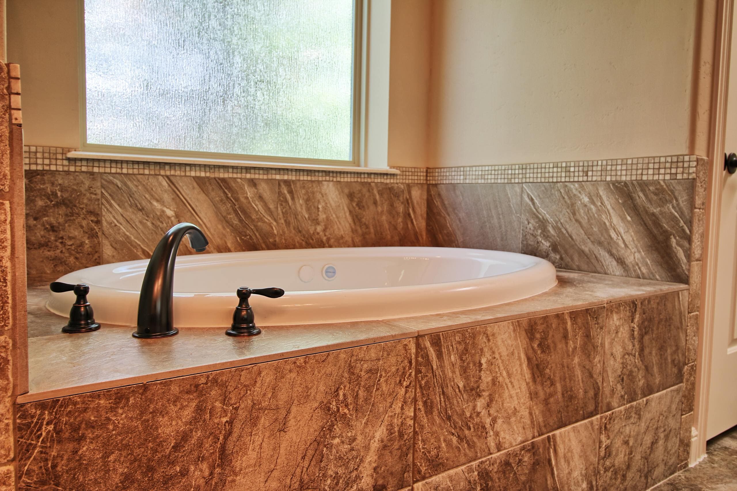 Drop-in jetted tub with oil-rubbed bronze faucet, brown marble-look tile surround and mosaic border below a textured glass window