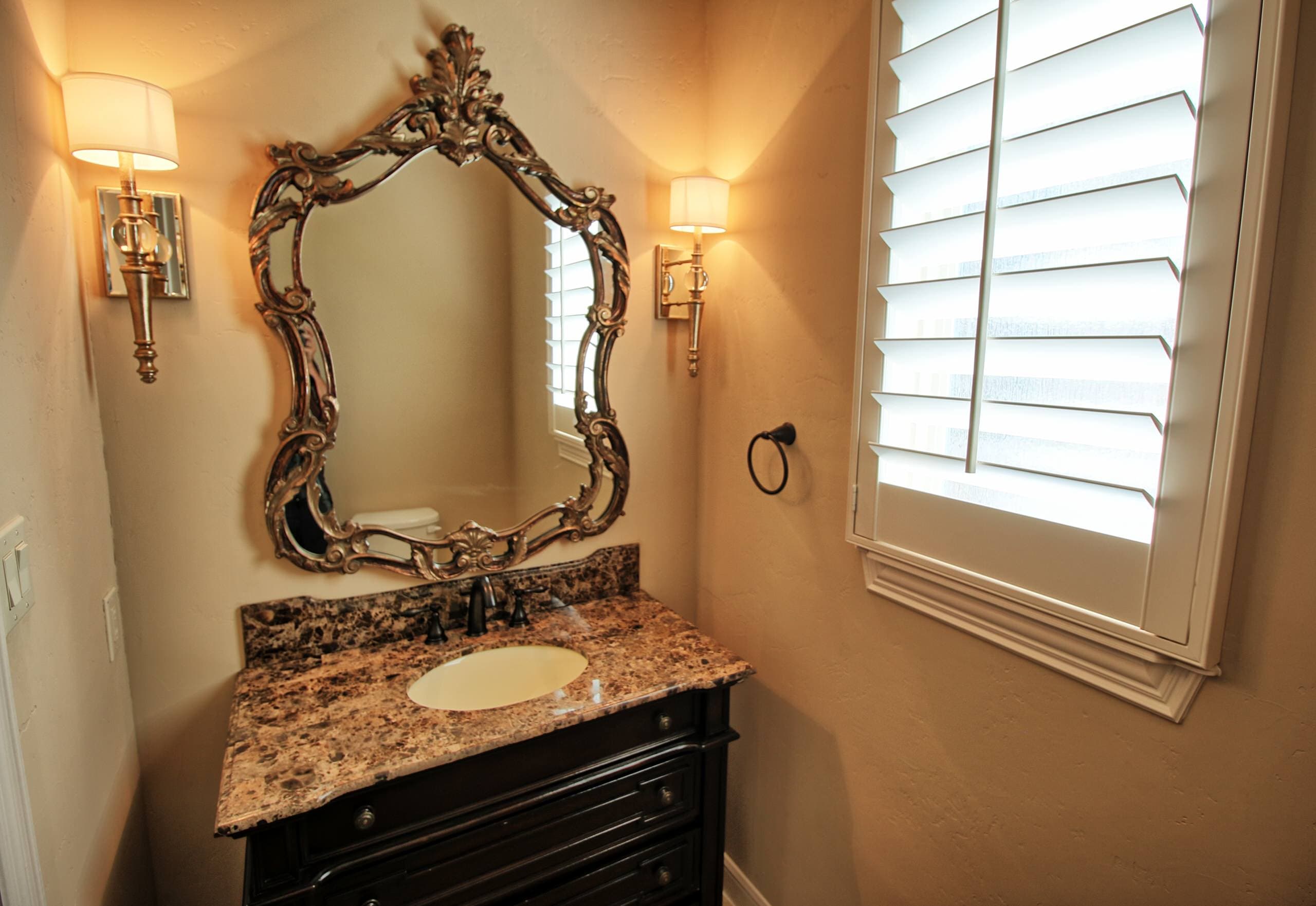 Powder room with dark furniture-style vanity, brown marble top, ornate gold-finish mirror, gold drum-shade sconces and white plantation shutters