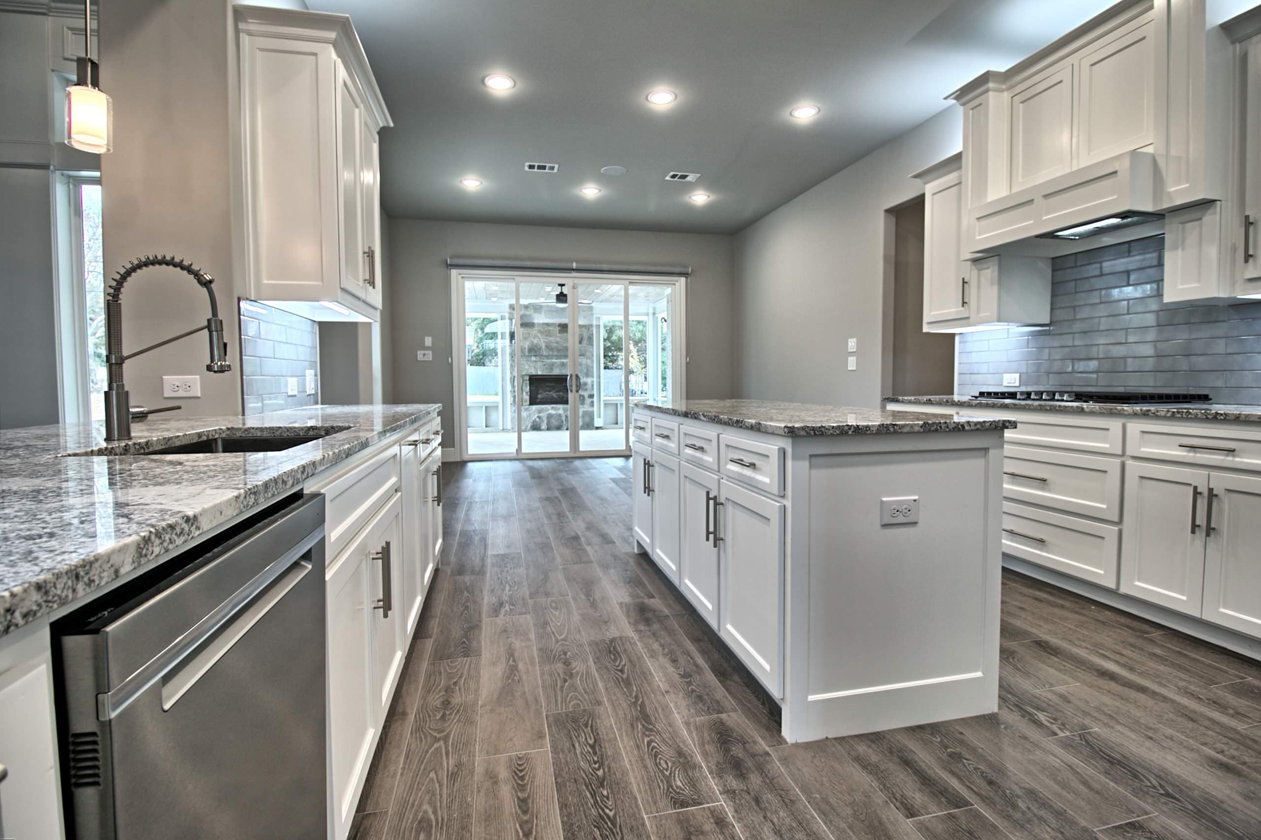 White shaker kitchen with island, granite counters, gray subway tile and wood-look floor; sliding doors reveal a covered patio with stone fireplace