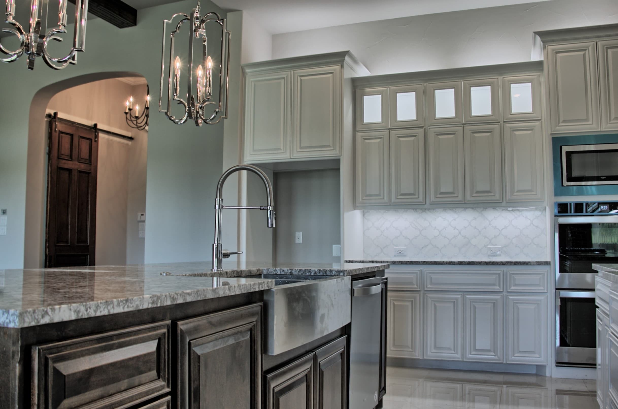 Dark stained island with granite top and stainless farmhouse sink, facing white raised-panel cabinets and an arch framing a barn door