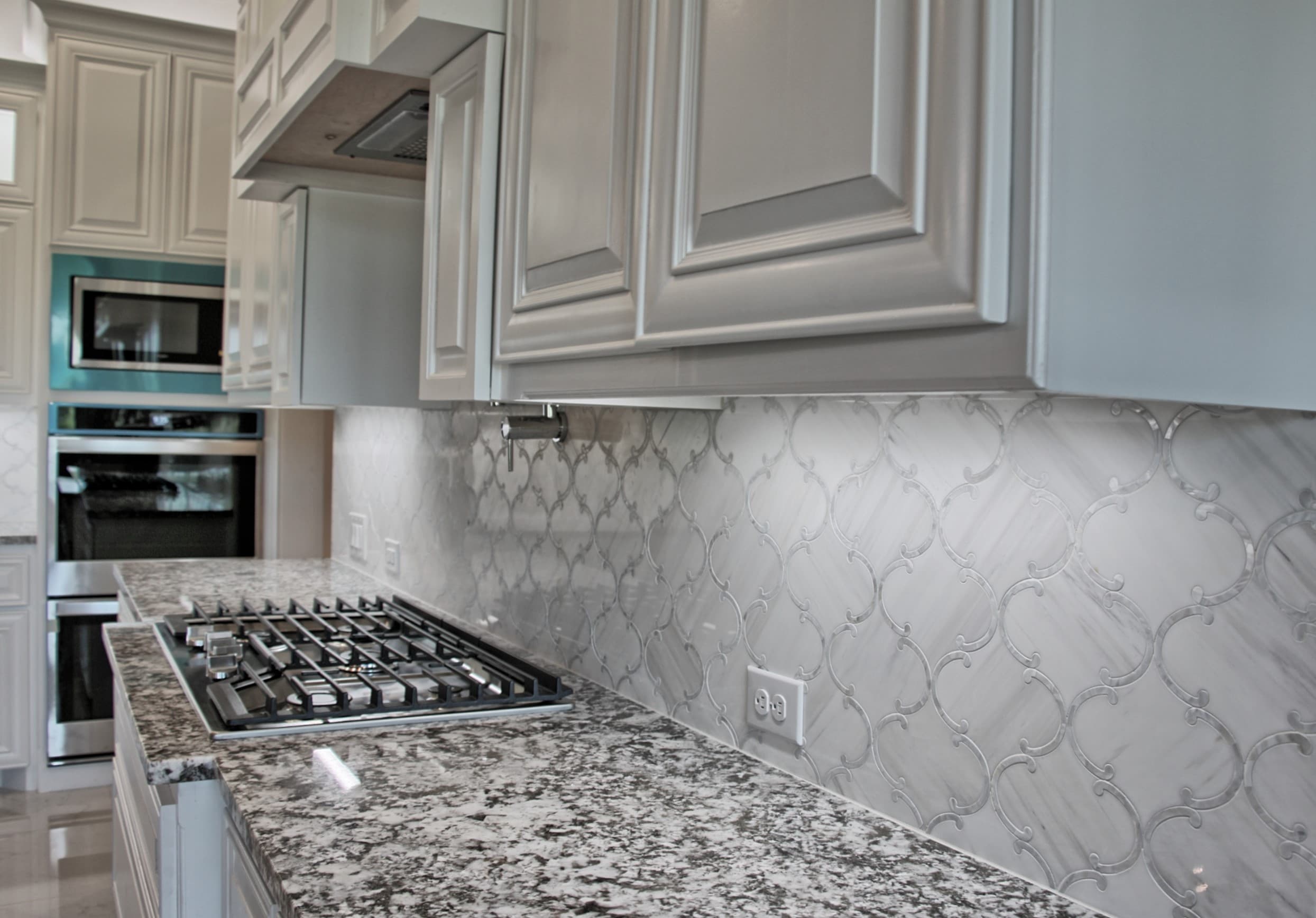Close view of a granite countertop with stainless gas cooktop, arabesque marble mosaic backsplash, pot filler and white raised-panel cabinets