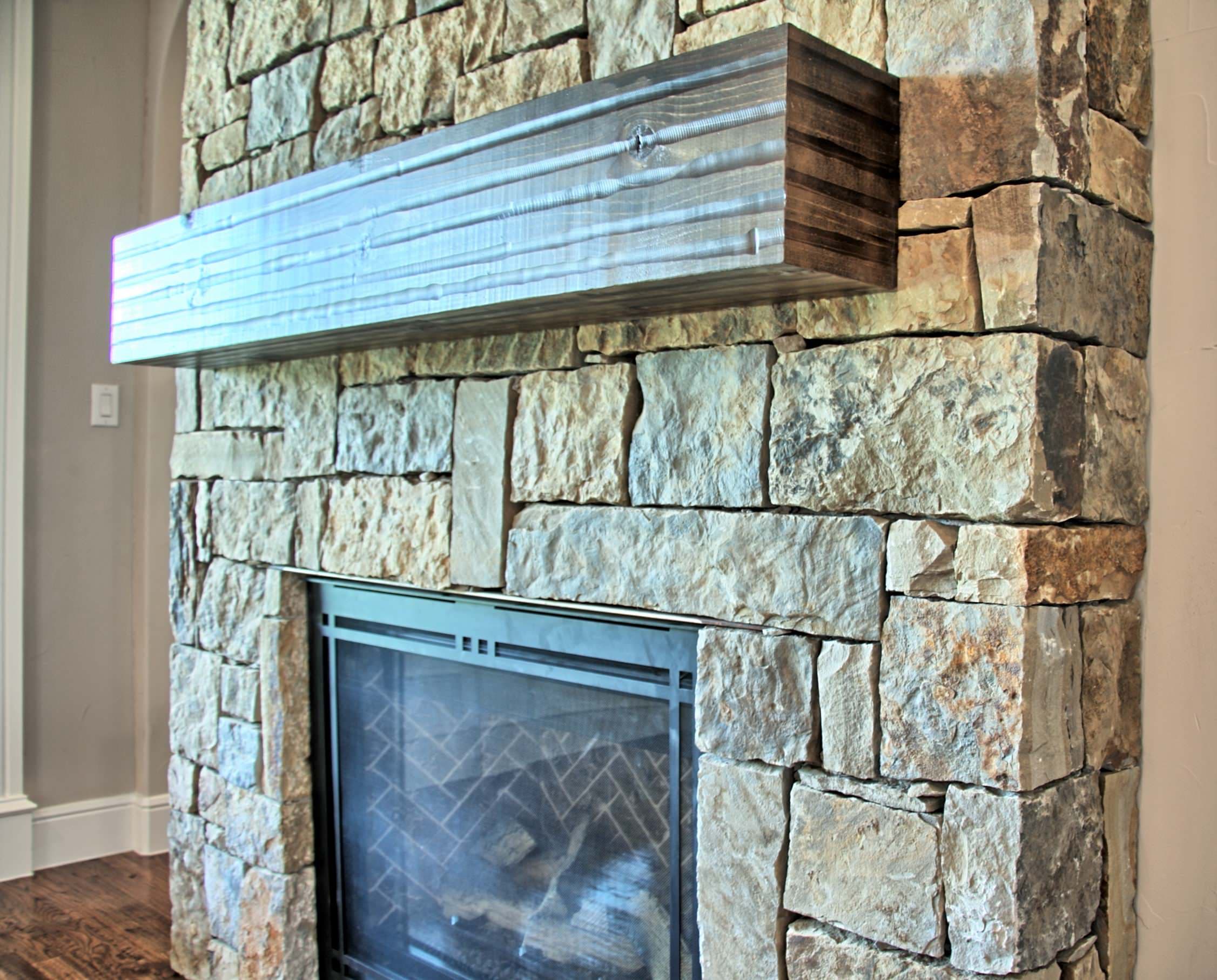 Close view of a stacked stone fireplace with a rough-sawn wood beam mantel and black metal screen over a herringbone-pattern firebox