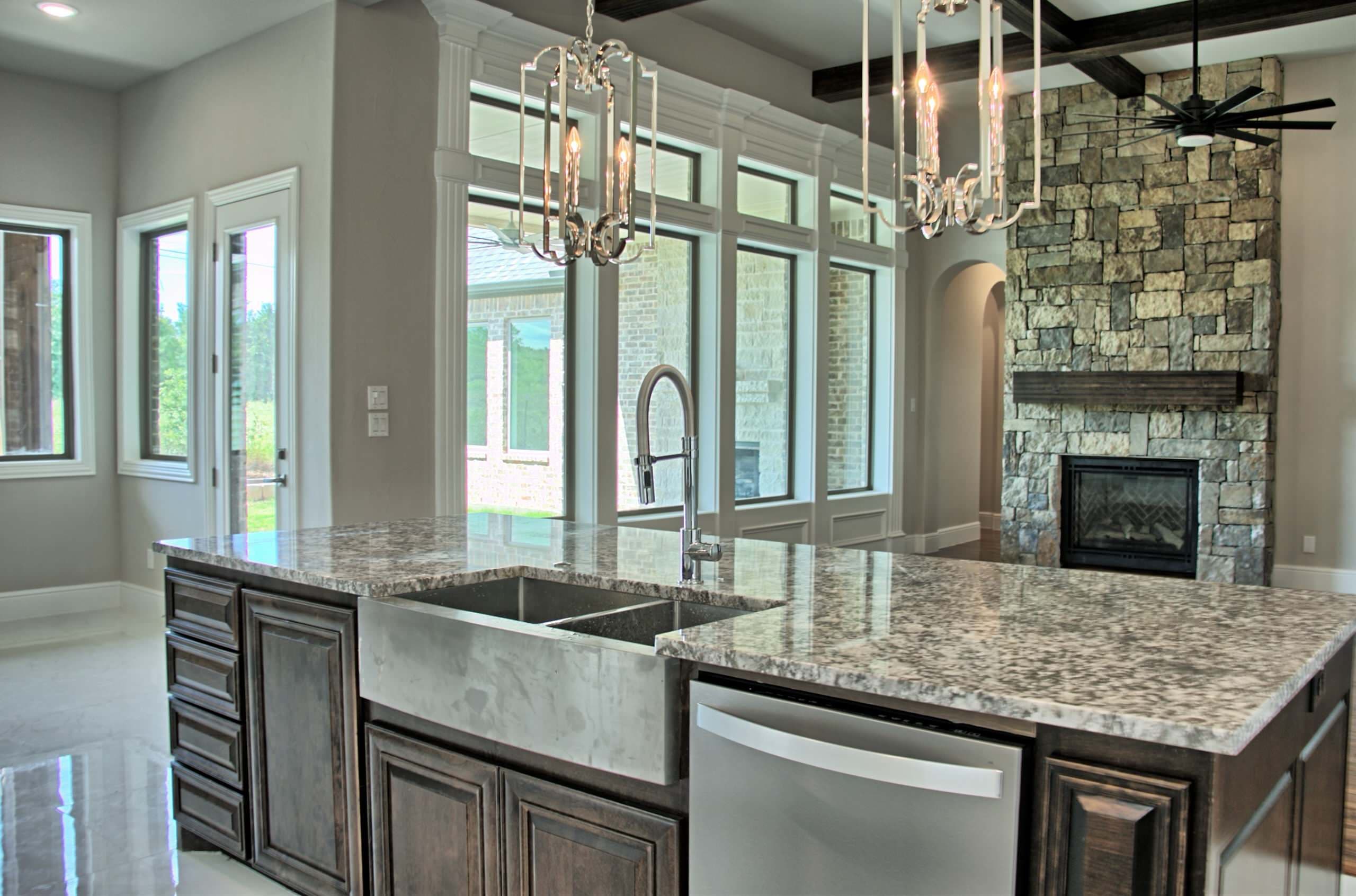 Dark stained island with granite top, farmhouse sink and chrome lantern pendants, facing a stacked stone fireplace under dark wood ceiling beams
