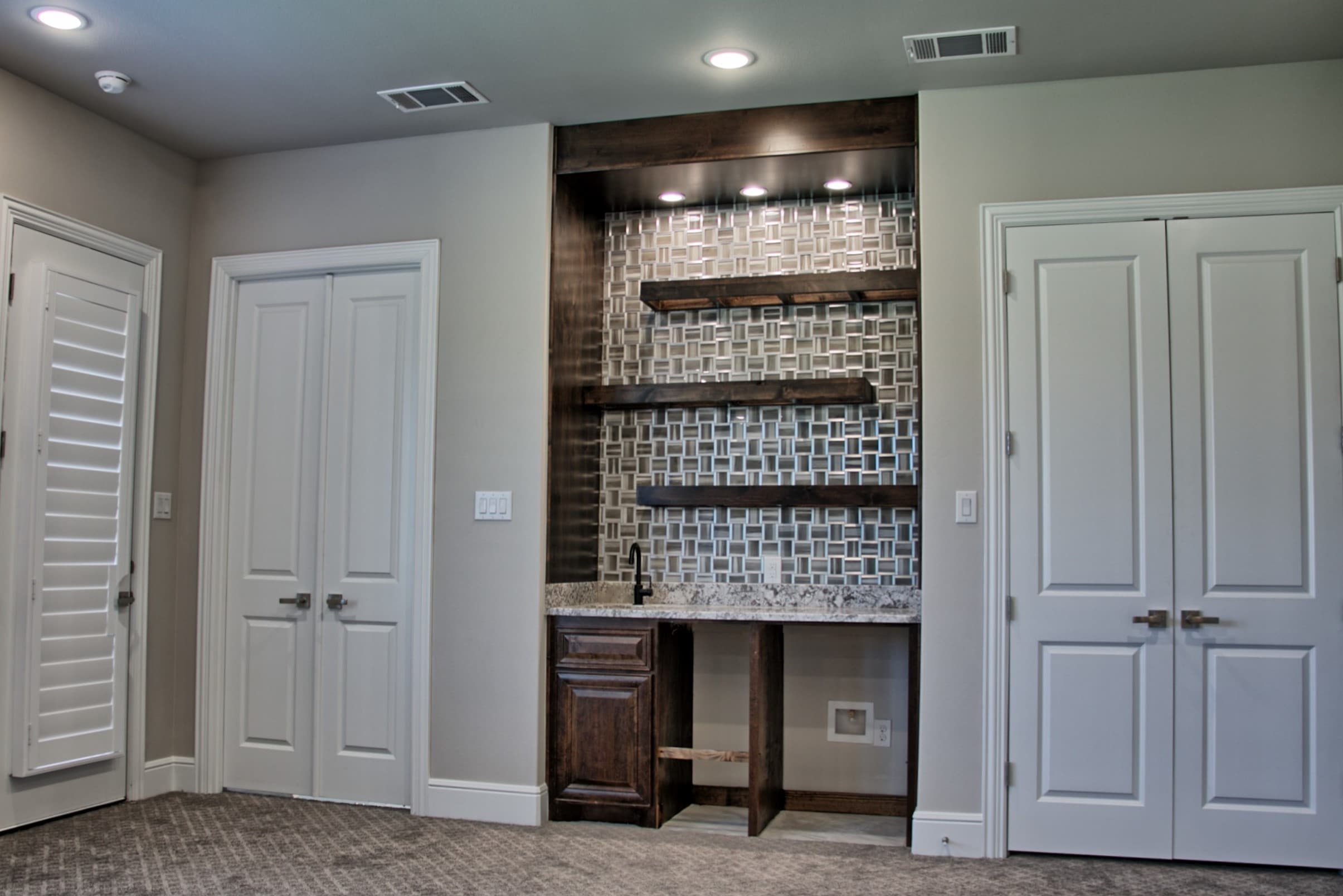 Built-in wet bar with dark stained wood surround, floating shelves, glass mosaic backsplash and granite counter, flanked by white double doors