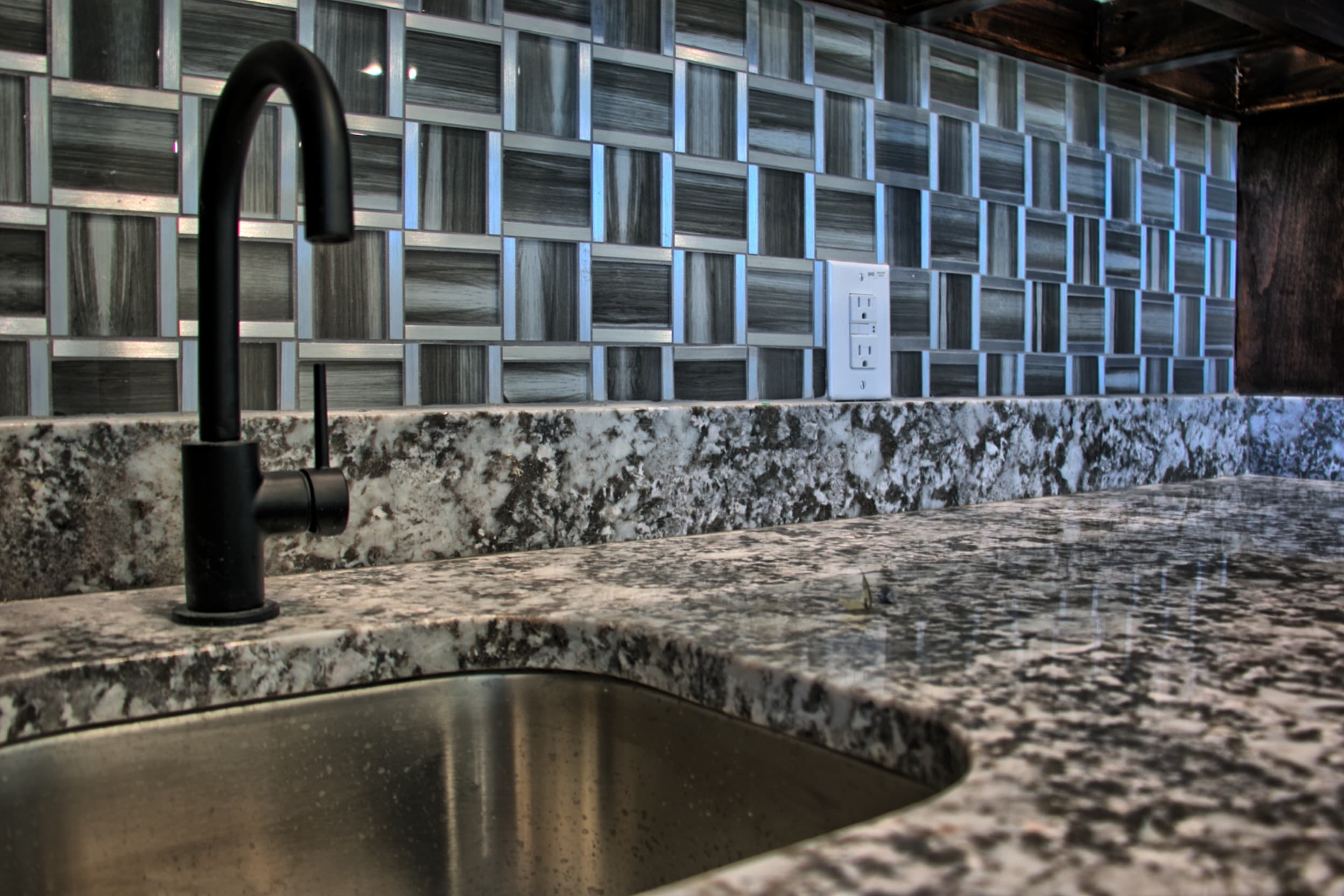 Matte black bar faucet and undermount stainless sink in a granite counter with a glass and metal basketweave mosaic backsplash